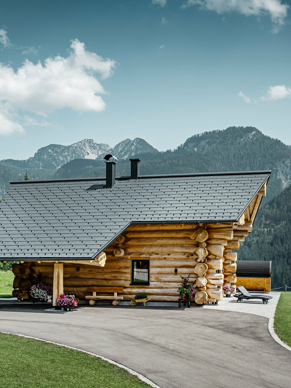 Blockhaus in Gosau, Österreich, mit einem PREFA Dachsystem in der Farbe Anthrazit. Die bewährten Dachplatten R.16 schaffen einen ansprechenden Kontrast zum natürlichen Holz des Blockhauses. Im Hintergrund erstreckt sich ein majestätisches Bergpanorama, das die ruhige und malerische Lage des Hauses unterstreicht. Die detailreiche Aufnahme fängt die Symbiose aus traditioneller Baukunst aus Österreich und moderner Dachtechnologie ein und zeigt die robuste und ästhetische Qualität der PREFA Aluminiumprodukte.