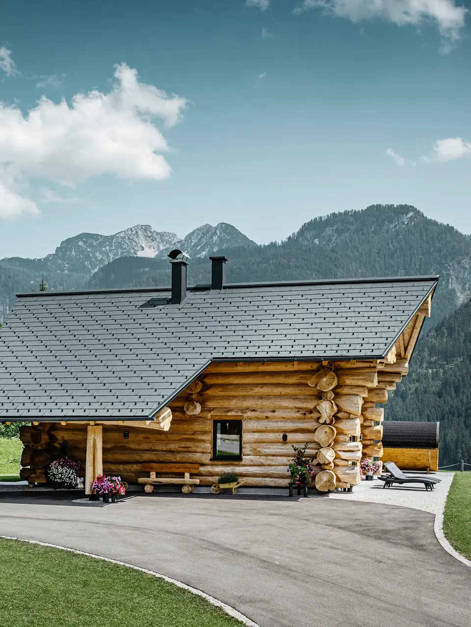 Blockhaus in Gosau, Österreich, mit einem PREFA Dachsystem in der Farbe Anthrazit. Die bewährten Dachplatten R.16 schaffen einen ansprechenden Kontrast zum natürlichen Holz des Blockhauses. Im Hintergrund erstreckt sich ein majestätisches Bergpanorama, das die ruhige und malerische Lage des Hauses unterstreicht. Die detailreiche Aufnahme fängt die Symbiose aus traditioneller Baukunst aus Österreich und moderner Dachtechnologie ein und zeigt die robuste und ästhetische Qualität der PREFA Aluminiumprodukte.