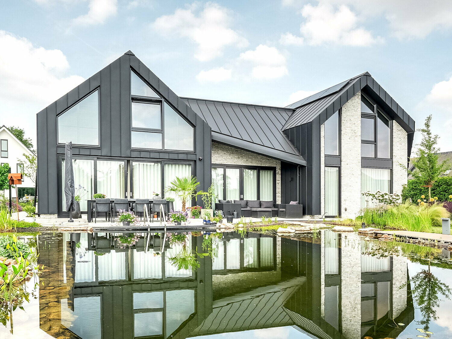 Modernes Einfamilienhaus mit charakteristischen, spitzen Giebeldächern, bedeckt mit schwarzen Prefalz-Dachbahnen, reflektiert sich in einem ruhigen Gartenteich. Die Fassade kombiniert dunkle Wandelemente mit hellen Ziegelbereichen und großen Fenstern. Eine Terrasse mit Gartenmöbeln lädt zum Verweilen ein und ist harmonisch in die üppige, blühende Gartenlandschaft integriert.