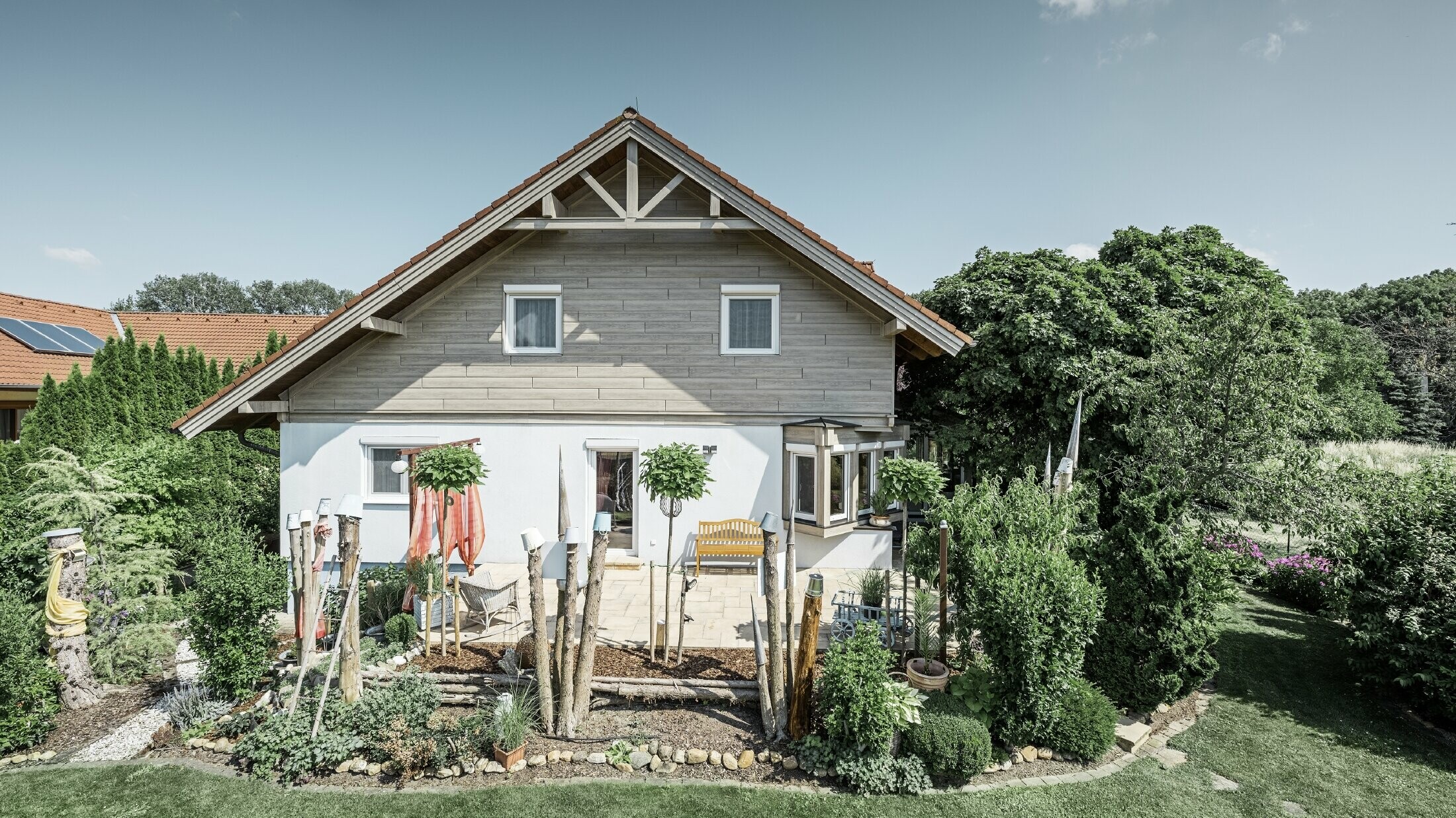 Fassadensaniertes Einfamilienhaus mit erneuerter Giebelverkleidung in moderner Holzoptik in Grau-Beige, kombiniert mit verputzter Fassade, Satteldach und gepflegtem Garten.