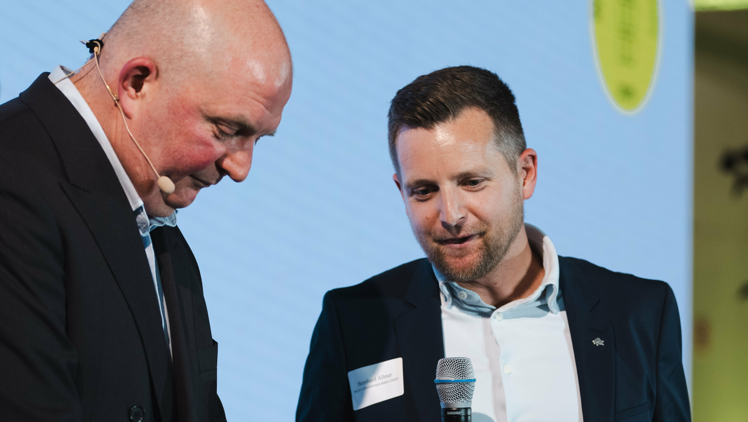 Preisübergabe beim Wettbewerb ‚Wohnbauten des Jahres‘ 2025: Zwei Personen auf der Bühne, eine hält ein Mikrofon und die Auszeichnung ‚Architects’ Choice‘, während die andere die Übergabe vorbereitet. Der Mann mit dem Mikrofon und der Auszeichnung ist Bernhard Allmer, der Produktverantwortliche für die PREFA Solarprodukte. Im Hintergrund leuchtet eine große Projektionsfläche mit Event-Logo und Akzenten in Grün. Die Szene steht für Innovation, Architekturqualität und die Auszeichnung des Solarmoduls PREFALZ als nachhaltige Lösung für moderne Dachsysteme.