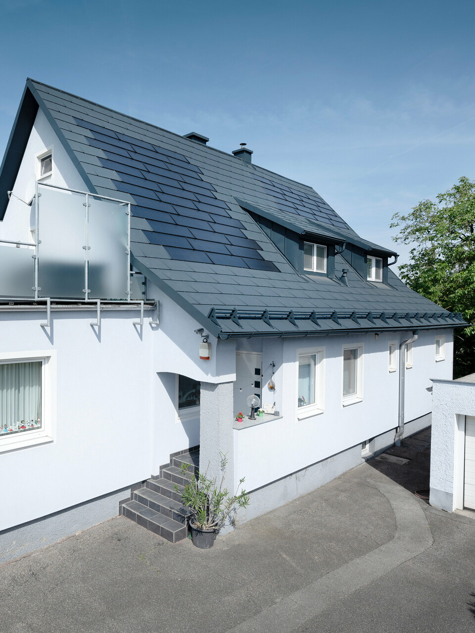 Dachsanierung eines Einfamilienhauses in Leonding mit PREFA Solardachplatten in Anthrazit. Das Bild zeigt das sanierte Haus aus einer seitlichen Perspektive mit klarer Sicht auf das Dach und die integrierten Solarmodule. Diese bieten eine nachhaltige und energieeffiziente Möglichkeit zur Stromerzeugung. Bei diesem Objekt erzeugen die Solardachplatten eine Stromleistung von 10,6 kWp. Die modernen, langlebigen und witterungsbeständigen PREFA Materialien tragen zu einer ästhetischen und funktionalen Dachkonstruktion bei, die den neuesten Standards für nachhaltiges Bauen entspricht.