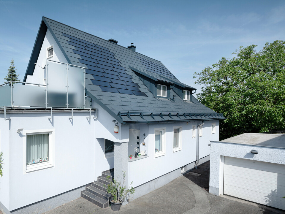 Dachsanierung eines Einfamilienhauses in Leonding mit PREFA Solardachplatten in Anthrazit. Das Bild zeigt das sanierte Haus aus einer seitlichen Perspektive mit klarer Sicht auf das Dach und die integrierten Solarmodule. Diese bieten eine nachhaltige und energieeffiziente Möglichkeit zur Stromerzeugung. Bei diesem Objekt erzeugen die Solardachplatten eine Stromleistung von 10,6 kWp. Die modernen, langlebigen und witterungsbeständigen PREFA Materialien tragen zu einer ästhetischen und funktionalen Dachkonstruktion bei, die den neuesten Standards für nachhaltiges Bauen entspricht.