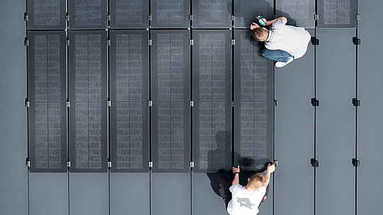 Zwei Personen installieren PREFALZ Solarmodule auf einer großen grauen Dachfläche, die mit Doppelstehfalzbahnen ausgelegt ist.