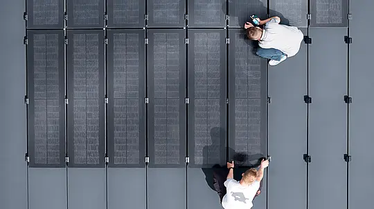 Zwei Personen installieren PREFALZ-Solarmodule auf einer großen grauen Dachfläche, die mit doppelflanschigen Paneelen bedeckt ist.