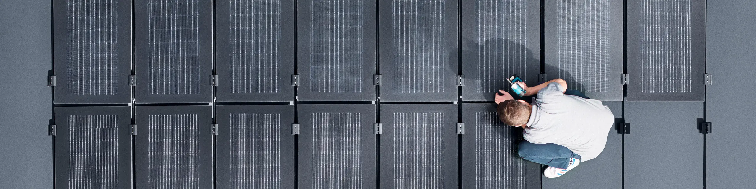 Zwei Personen installieren PREFALZ-Solarmodule auf einer großen grauen Dachfläche, die mit doppelflanschigen Paneelen bedeckt ist.