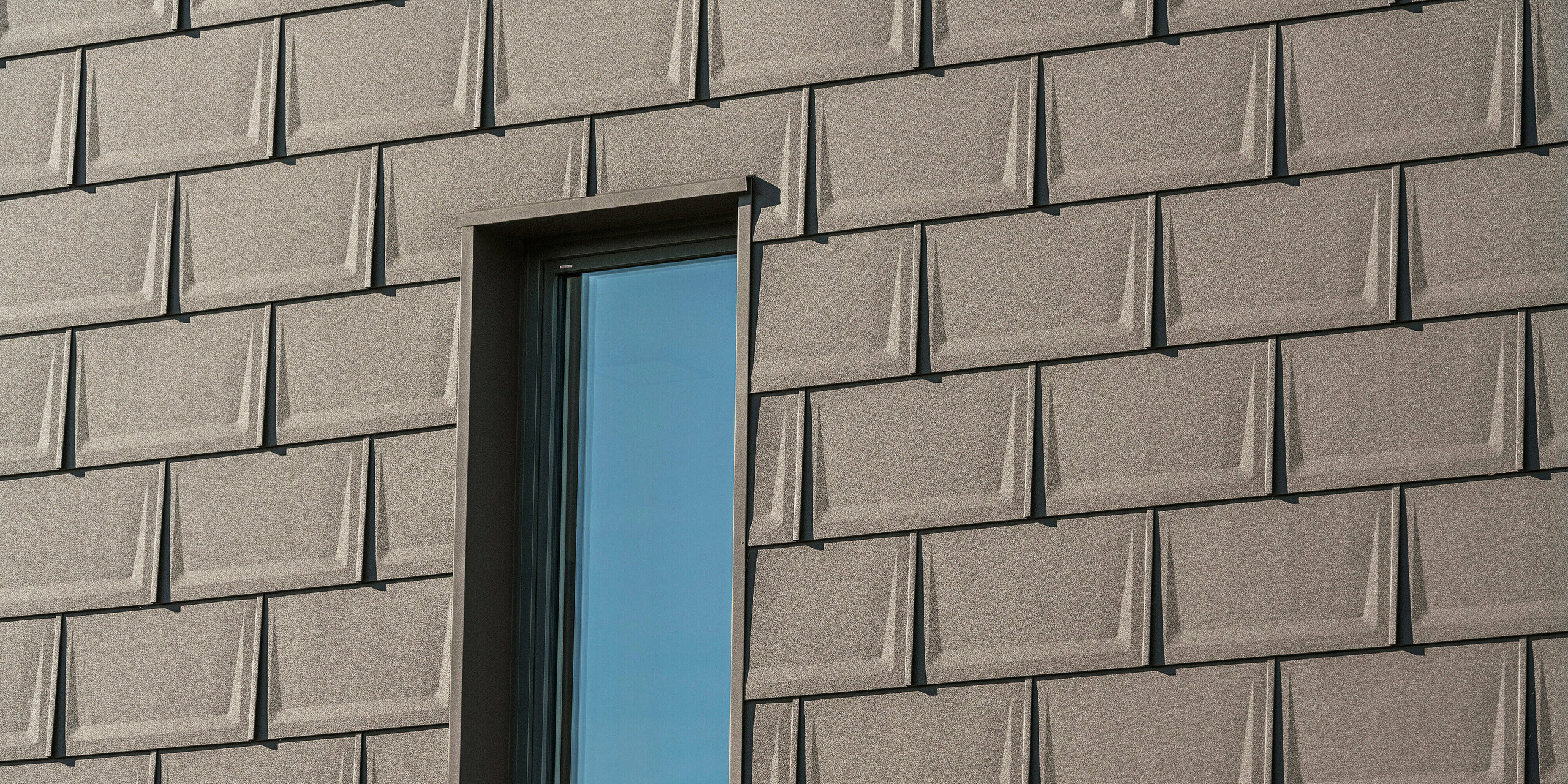 Detailaufnahme eines Fensters an der Fassade eines Einfamilienhauses in Neukirchen, Österreich, akzentuiert durch die PREFA Dachplatte R.16 in P.10 Braun. Die einheitliche Verlegung der Platten schafft eine dynamische Textur und verleiht der Hausfassade einen modernen Charakter. Die Präzision der Installation und die harmonische Farbabstimmung unterstreichen das qualitätsbewusste Baudesign, das für PREFA Aluminiumprodukte steht.