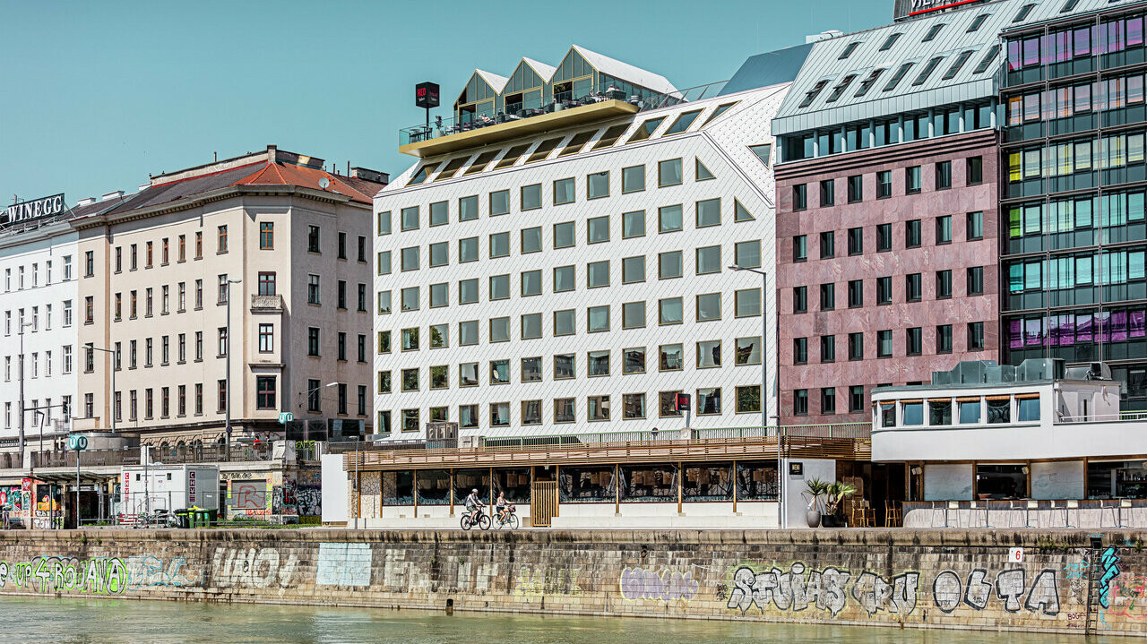 Blick auf das Ufer des Donaukanals in Wien mit einer Reihe von Gebäuden unterschiedlicher Architektur. Im Vordergrund ist das "THE ROCK Radisson RED Vienna" Hotel, erkennbar an seiner charakteristischen weißen PREFA Aluminiumfassade mit Dach- und Wandrauten 44 × 44 in P.10 Reinweiß.