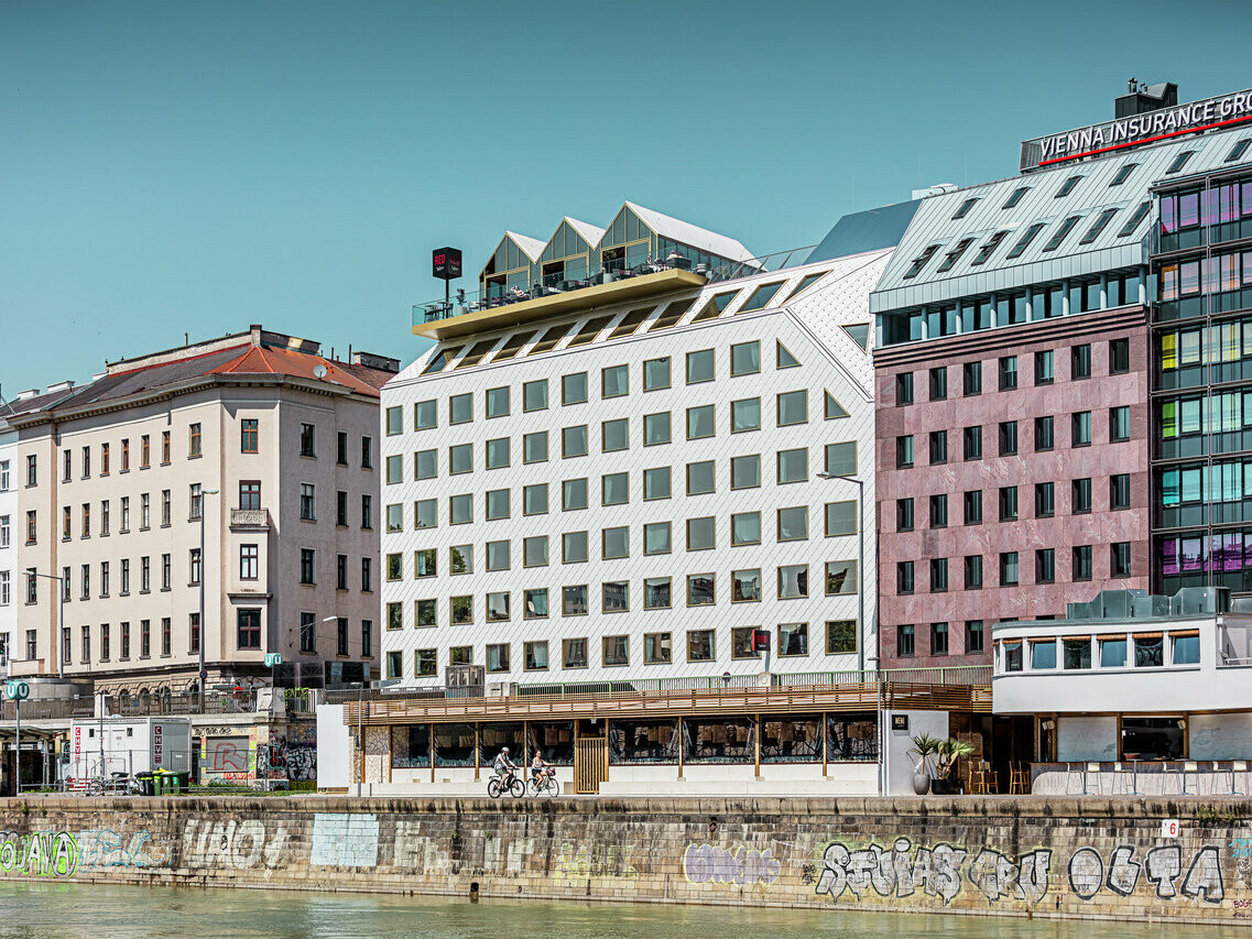 Blick auf das Ufer des Donaukanals in Wien mit einer Reihe von Gebäuden unterschiedlicher Architektur. Im Vordergrund ist das "THE ROCK Radisson RED Vienna" Hotel, erkennbar an seiner charakteristischen weißen PREFA Aluminiumfassade mit Dach- und Wandrauten 44 × 44 in P.10 Reinweiß.