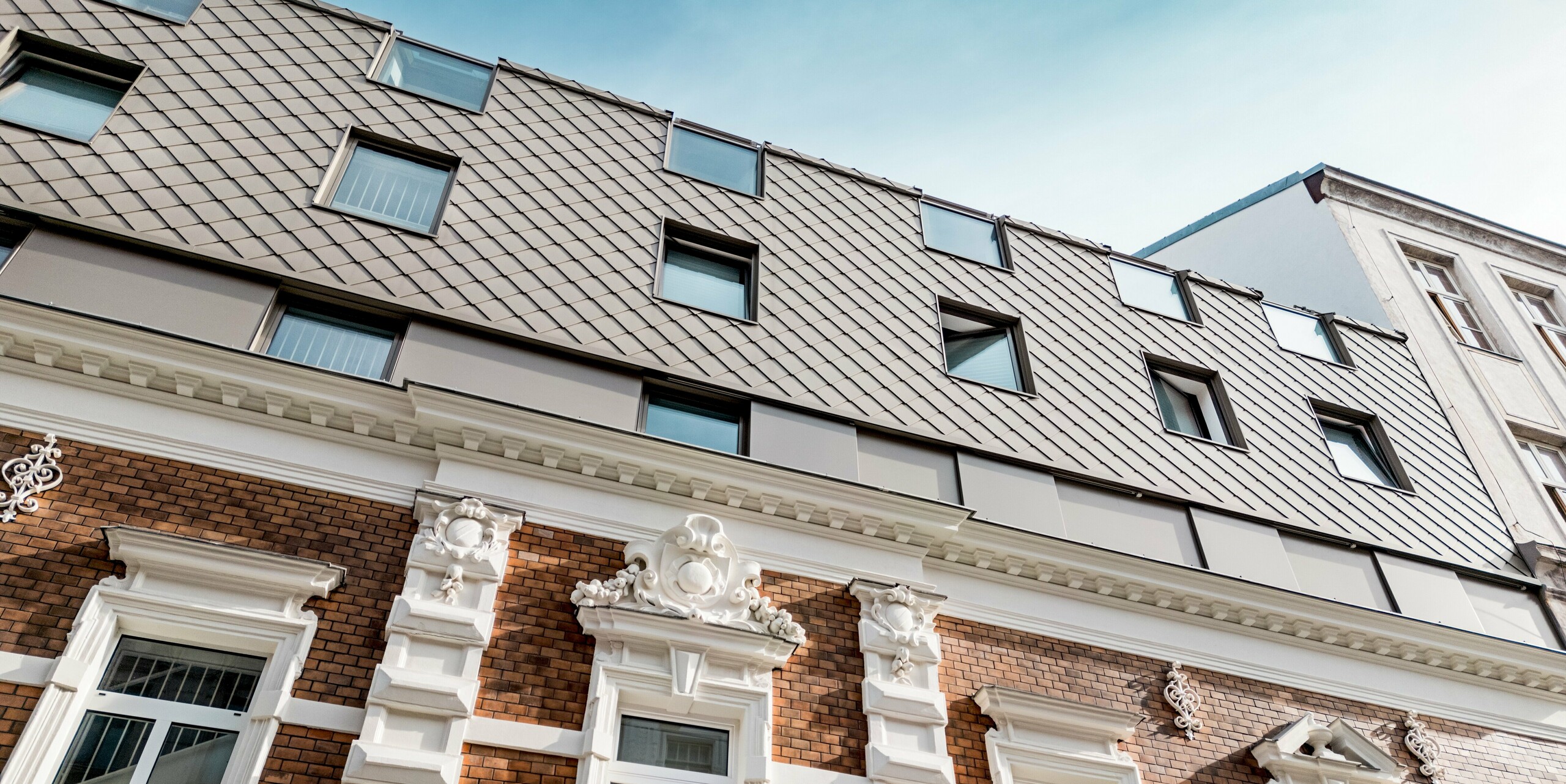 Das Bild zeigt die gelungene Verbindung von historischer und moderner Architektur in einem Wiener Wohngebäude. Die klassischen Elemente der Fassade mit prachtvollen Stuckarbeiten und roten Ziegeln werden durch die modernen PREFA Rauten 29×29 in Bronze auf dem Dachbereich stilvoll ergänzt. Die präzise angeordneten Fenster in der oberen Etage fügen sich harmonisch in das Gesamtbild ein, während die Dachkonstruktion mit den Rauten eine zeitgemäße Note verleiht. Diese Kombination von traditioneller und innovativer Gestaltung verleiht dem Gebäude ein einzigartiges, ästhetisches Erscheinungsbild.