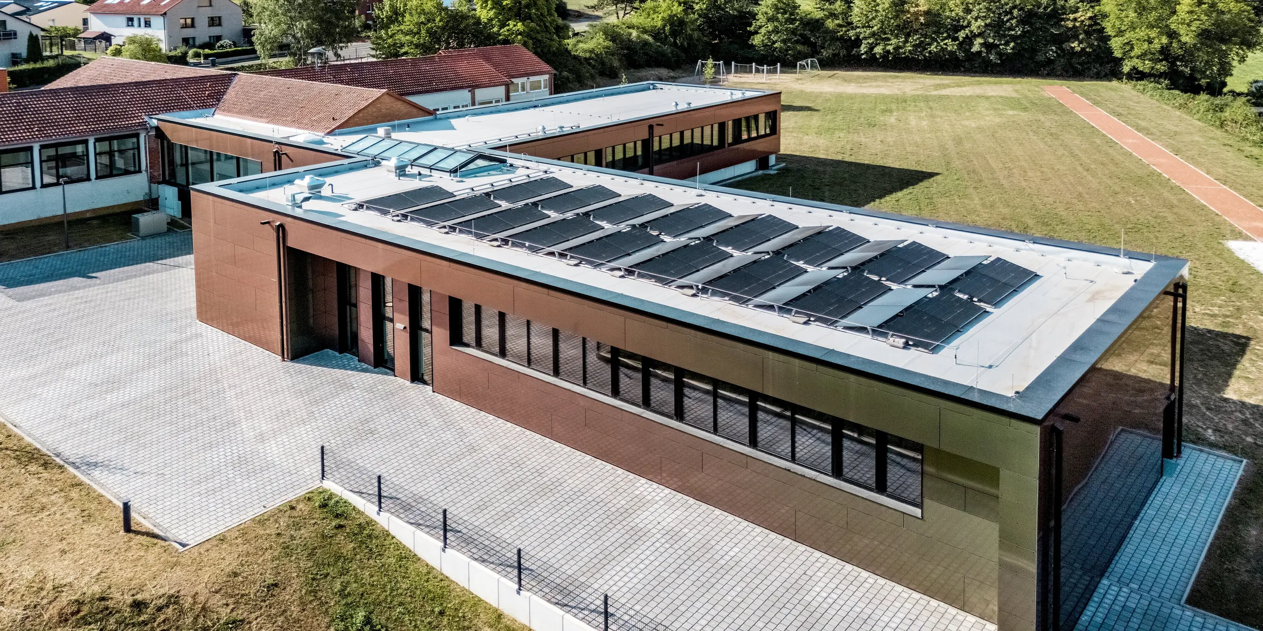 Beeindruckende Vogelperspektive auf die Johannwolfschule Angerstein: Die modernen, flachen Gebäudetrakte mit großzügigen Fensterfronten und Photovoltaikanlage fügen sich harmonisch in die grüne Landschaft ein. Die PREFA Aluminium Verbundplatten in der Sonderfarbe Spring Lake schimmern in warmen Bronze- und Grüntönen. Dunkle PREFA Quadratrohre ergänzen die langlebige, witterungsbeständige Fassadengestaltung.