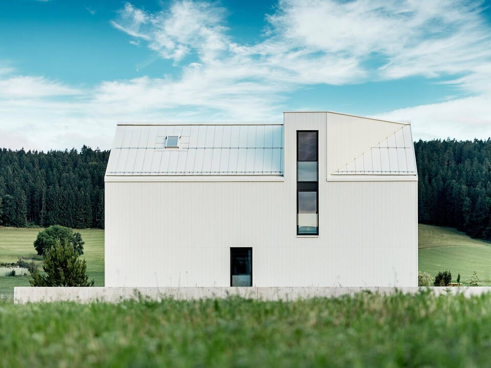 Das Bild zeigt das Einfamilienhaus in Freistadt aus einer Seitenansicht, die die markante, minimalistische Architektur des Gebäudes hervorhebt. Die Fassade aus reinweißem PREFALZ-Aluminium und das steile Satteldach schaffen eine klare, moderne Silhouette, die sich harmonisch in die grüne Umgebung einfügt. Die großen Fensterflächen und das elegante Gaubenfenster betonen das funktionale Design, während das hochwertige Aluminiummaterial sowohl ästhetische Beständigkeit als auch Witterungsresistenz gewährleistet. Das Gebäude strahlt durch seine einfache Form und die Verwendung langlebiger Materialien eine zeitlose Eleganz aus und stellt eine perfekte Kombination aus Architektur und Naturverbundenheit dar.