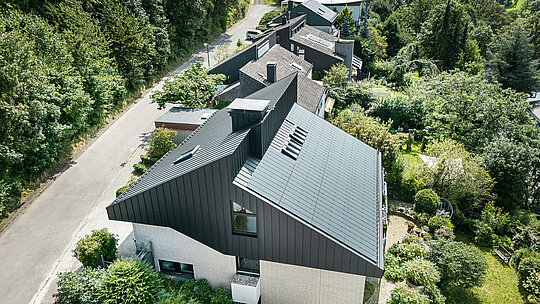 Schrägansicht auf ein modernes Einfamilienhaus in Meschede, Deutschland. Das versetzte Pultdach wurde mit PREFALZ in P.10 Schwarz verkleidet. Das Aluminiumdach dient als optisches Highlight und bietet dem Gebäude dauerhaften Schutz. Die präzise Doppelstehfalzdeckung zieht sich vom Dach über die Fassade an den Giebelseiten. Umgeben von üppigem Grün und einer ruhigen Straße fügt sich das energieeffiziente Gebäude mit den innovativen PREFALZ Solarmodulen perfekt in das naturnahe Wohnumfeld ein.