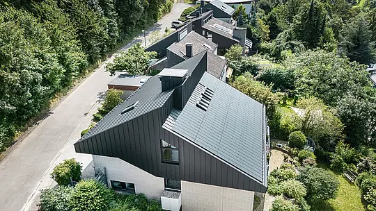 Schrägansicht eines modernen Einfamilienhauses in Meschede, Deutschland. Das Satteldach ist mit PREFALZ P.10 Schwarz gedeckt. Das Aluminiumdach setzt einen optischen Akzent und bietet dem Gebäude langfristigen Schutz. Die präzise Doppeldachkonstruktion erstreckt sich vom Dach über die Fassade bis zur Vorderseite. Umgeben von üppigem Grün und einer ruhigen Straße fügt sich das energieeffiziente Haus mit innovativen PREFALZ-Solarmodulen harmonisch in die natürliche Umgebung ein.