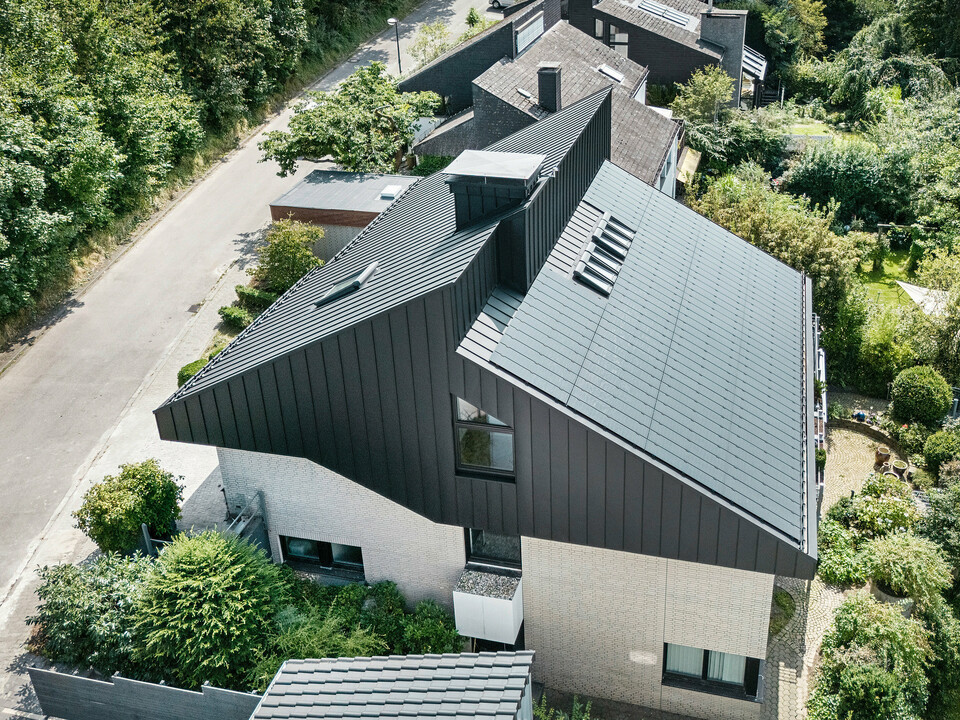 Schrägansicht auf ein modernes Einfamilienhaus in Meschede, Deutschland. Das versetzte Pultdach wurde mit PREFALZ in P.10 Schwarz verkleidet. Das Aluminiumdach dient als optisches Highlight und bietet dem Gebäude dauerhaften Schutz. Die präzise Doppelstehfalzdeckung zieht sich vom Dach über die Fassade an den Giebelseiten. Umgeben von üppigem Grün und einer ruhigen Straße fügt sich das energieeffiziente Gebäude mit den innovativen PREFALZ Solarmodulen perfekt in das naturnahe Wohnumfeld ein.