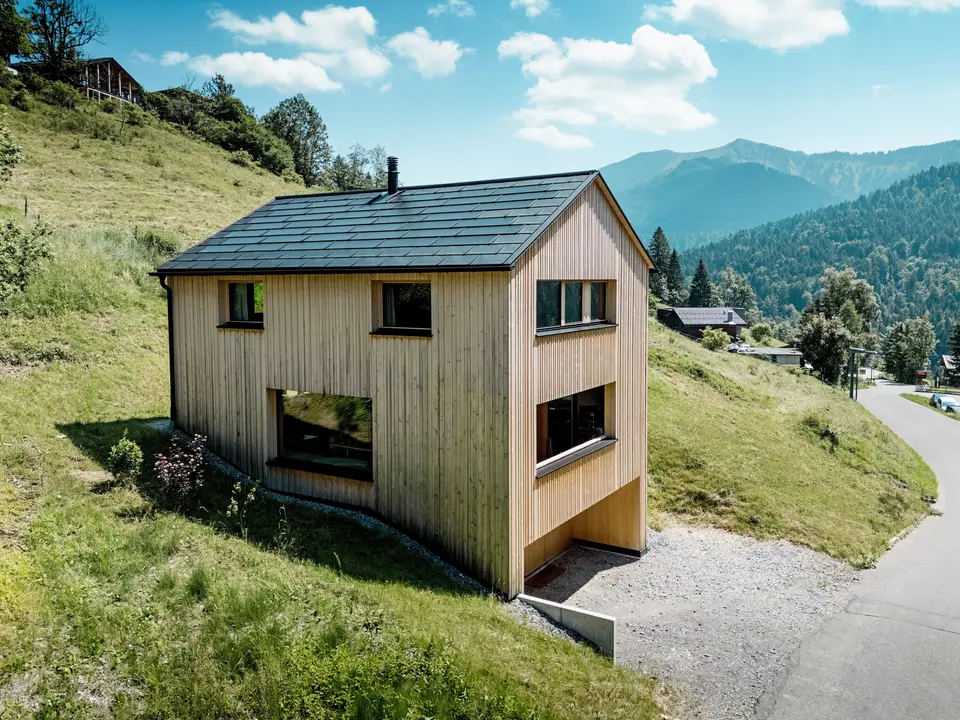 Hoch über dem Tal steht Haus Immi wie ein klarer Holzmonolith im sonnigen Berghang von Ebnit. Die reduzierte Weißtannenfassade und das schwarze PREFA Solardach setzen einen kraftvollen Kontrast zur weiten Alpenlandschaft. Integrierte Solardachplatten und Dachplatten R.16 in P.10 Schwarz liefern Solarstrom, trotzen alpinem Wetter und sichern ein langlebiges, harmonisches Dachbild.