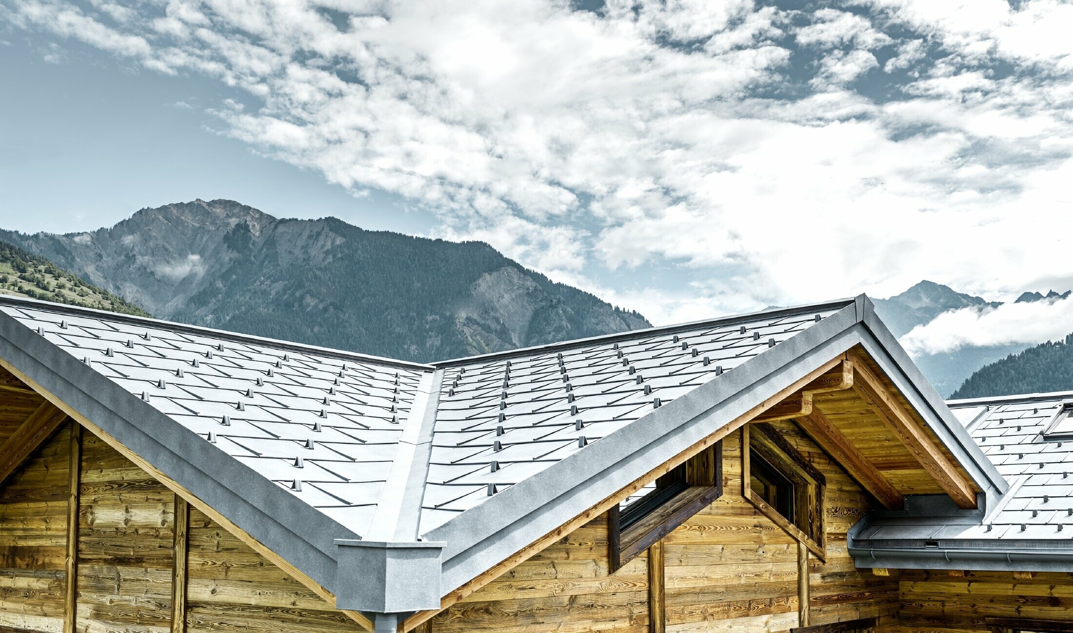 Dach einer rustikalen Hütte in der Schweiz mit Holzfassade und einem Aludach von PREFA. Verlegt wurde die Dachplatte R.16 in Steingrau. Man erkennt das verwinkelte Dach und die Kehle.