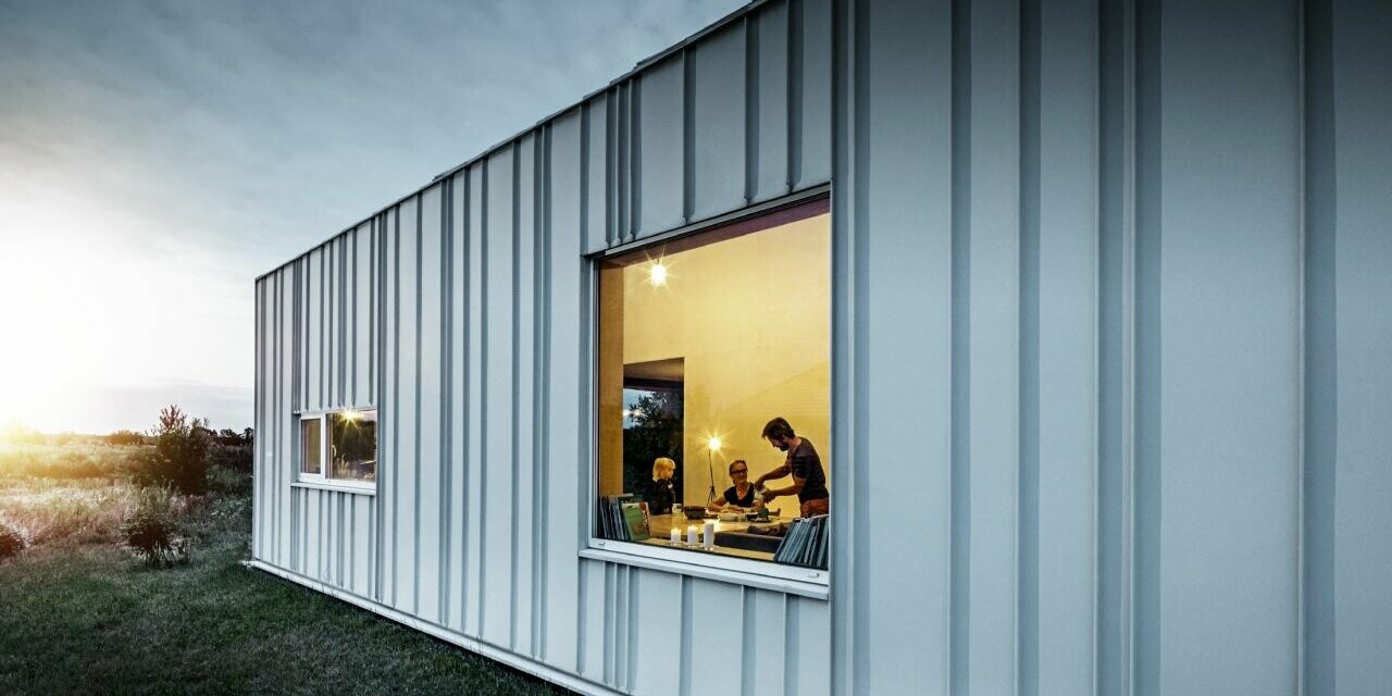 Winkelstehfalz Fassade mit PREFALZ in unterschiedlichen Scharenbreiten in P.10 Prefaweiß bei Dämmerung fotografiert. Durch das Esszimmer erkennt man eine Familie beim Abendessen. 