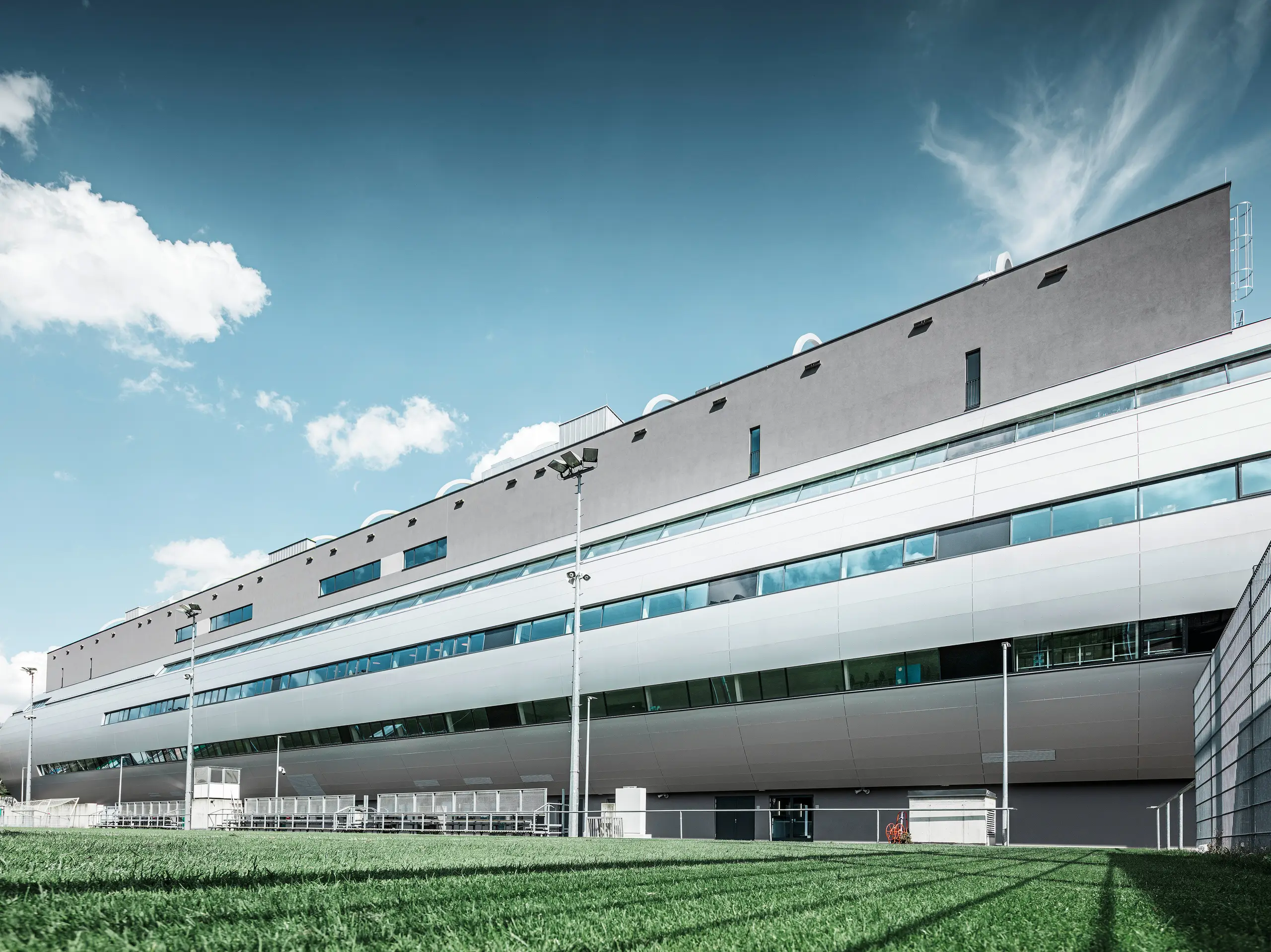 Das Allianz Stadion von SK Rapid Wien, abgebildet vom Trainingsplatz, zeigt die glänzende Fassade aus PREFA Aluminium Verbundplatten in Silbermetallic. Die harmonische Verschmelzung von funktionaler Sportstätte und eleganter Architektur wird durch die Fensterreihen betont. Die grüne Wiese des Trainingsplatzes am unteren Bildrand wird durch die Betreuerbänke und den Zaun abgegrenzt. Am Zaus sind zudem hohe Flutlichtmasten zu erkennen, die eine Ausübung des Trainings zu jeder Tageszeit ermöglichen. Der blaue Himmel komplementiert das professionelle Foto.