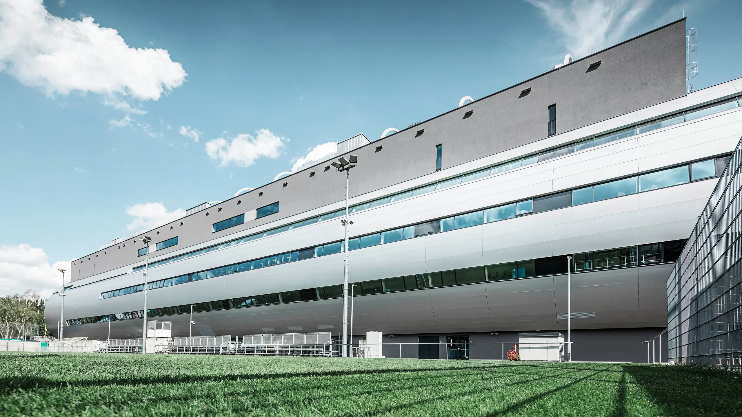 Das Allianz Stadion von SK Rapid Wien, abgebildet vom Trainingsplatz, zeigt die glänzende Fassade aus PREFA Aluminium Verbundplatten in Silbermetallic. Die harmonische Verschmelzung von funktionaler Sportstätte und eleganter Architektur wird durch die Fensterreihen betont. Die grüne Wiese des Trainingsplatzes am unteren Bildrand wird durch die Betreuerbänke und den Zaun abgegrenzt. Am Zaus sind zudem hohe Flutlichtmasten zu erkennen, die eine Ausübung des Trainings zu jeder Tageszeit ermöglichen. Der blaue Himmel komplementiert das professionelle Foto.