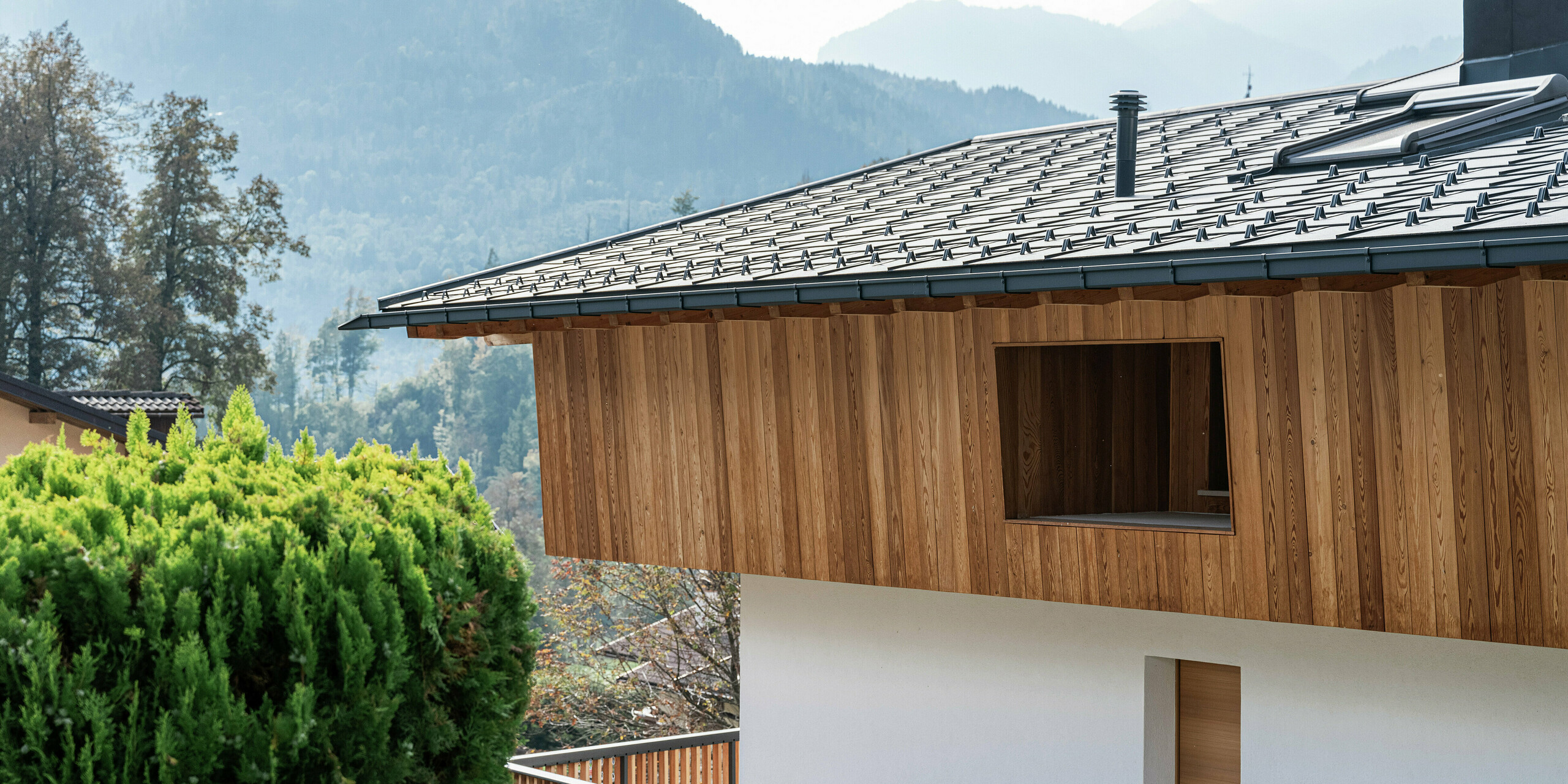 Detailansicht eines traditionellen Einfamilienhauses in Agordo, Italien, mit einer natürlichen Holzfassade und robusten PREFA Dachplatten R.16 in P.10 Anthrazit. Das widerstandsfähige Aluminiumdach mit Schneestoppern sorgt für optimalen Witterungsschutz. Die Kombination aus naturbelassener Holzfassade im Obergeschoss und weißer Putzfassade im Erdgeschoss verleiht dem Haus ein warmes, modernes Erscheinungsbild. Die klare Linienführung und die hochwertigen Materialien fügen sich harmonisch in die alpine Umgebung ein.