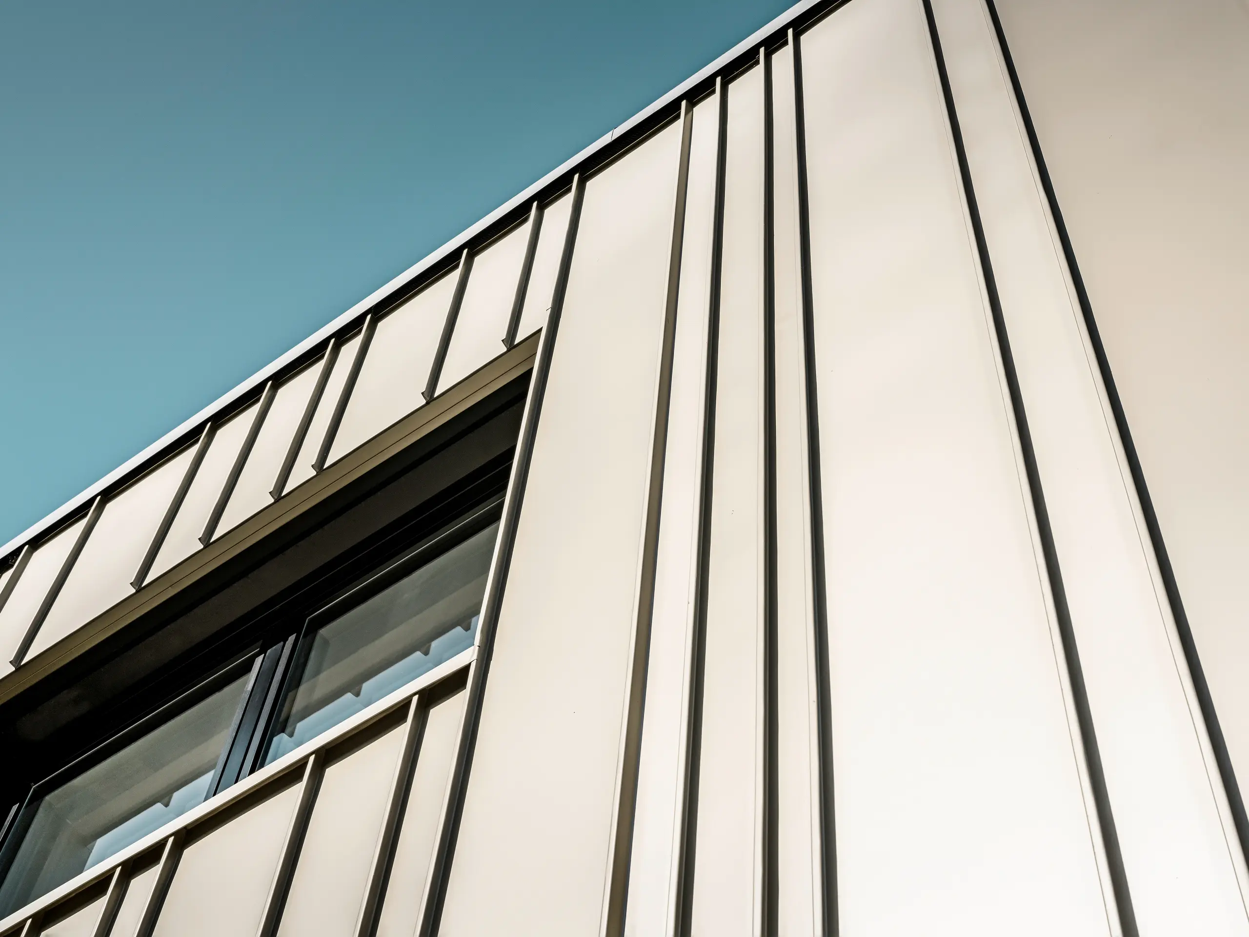 Nahaufnahme einer modernen PREFA Aluminiumfassade in Lichtbronze. Für die Winkelstehfalzfassade wurde FALZONAL in unterschiedlichen Scharenbreiten verlegt. Die Schatten der vertikalen Linien, die durch die Winkelstehfalzdeckung entstehen, heben das anspruchsvolle Design und die Präzision der Verarbeitung hervor. Das Fenster mit seiner schmalen, anthrazitfarbenen Einfassung fügt sich nahtlos in die Struktur der Fassade ein und spiegelt die Kombination aus Funktionalität, moderner Ästhetik und technischer Perfektion wider.