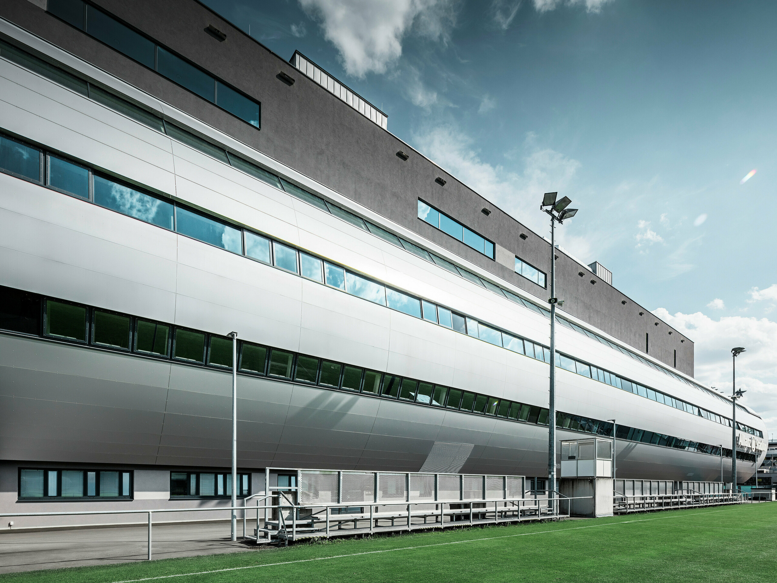 Seitenansicht des Allianz Stadions von SK Rapid Wien, charakterisiert durch die elegante PREFA Fassade in Form von Aluminium Verbundplatten in Silbermetallic. Die Architektur kombiniert Form und Funktion, mit langgezogenen Fensterfronten, die einen Blick in das sportliche Treiben gewähren und das Stadion zu einem Symbol für zeitgenössisches Design und sportliche Exzellenz machen. Der Blick vom Trainingsgelände zeigt den Blick der Spieler auf den gepflegten Rasen, die hohen Flutlichtmasten sowie die bequemen Betreuerbänke.