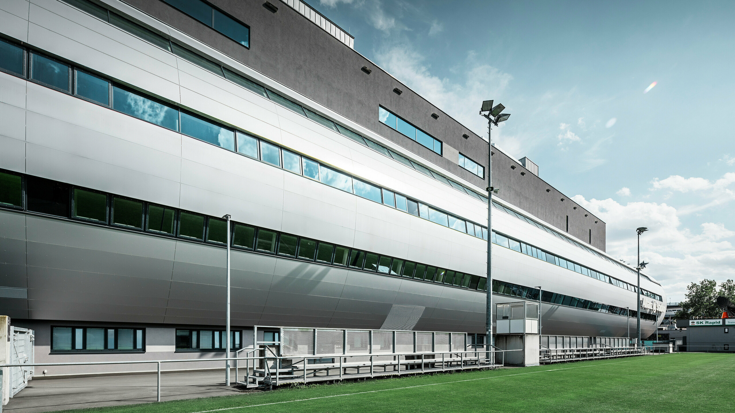 Seitenansicht des Allianz Stadions von SK Rapid Wien, charakterisiert durch die elegante PREFA Fassade in Form von Aluminium Verbundplatten in Silbermetallic. Die Architektur kombiniert Form und Funktion, mit langgezogenen Fensterfronten, die einen Blick in das sportliche Treiben gewähren und das Stadion zu einem Symbol für zeitgenössisches Design und sportliche Exzellenz machen. Der Blick vom Trainingsgelände zeigt den Blick der Spieler auf den gepflegten Rasen, die hohen Flutlichtmasten sowie die bequemen Betreuerbänke. 