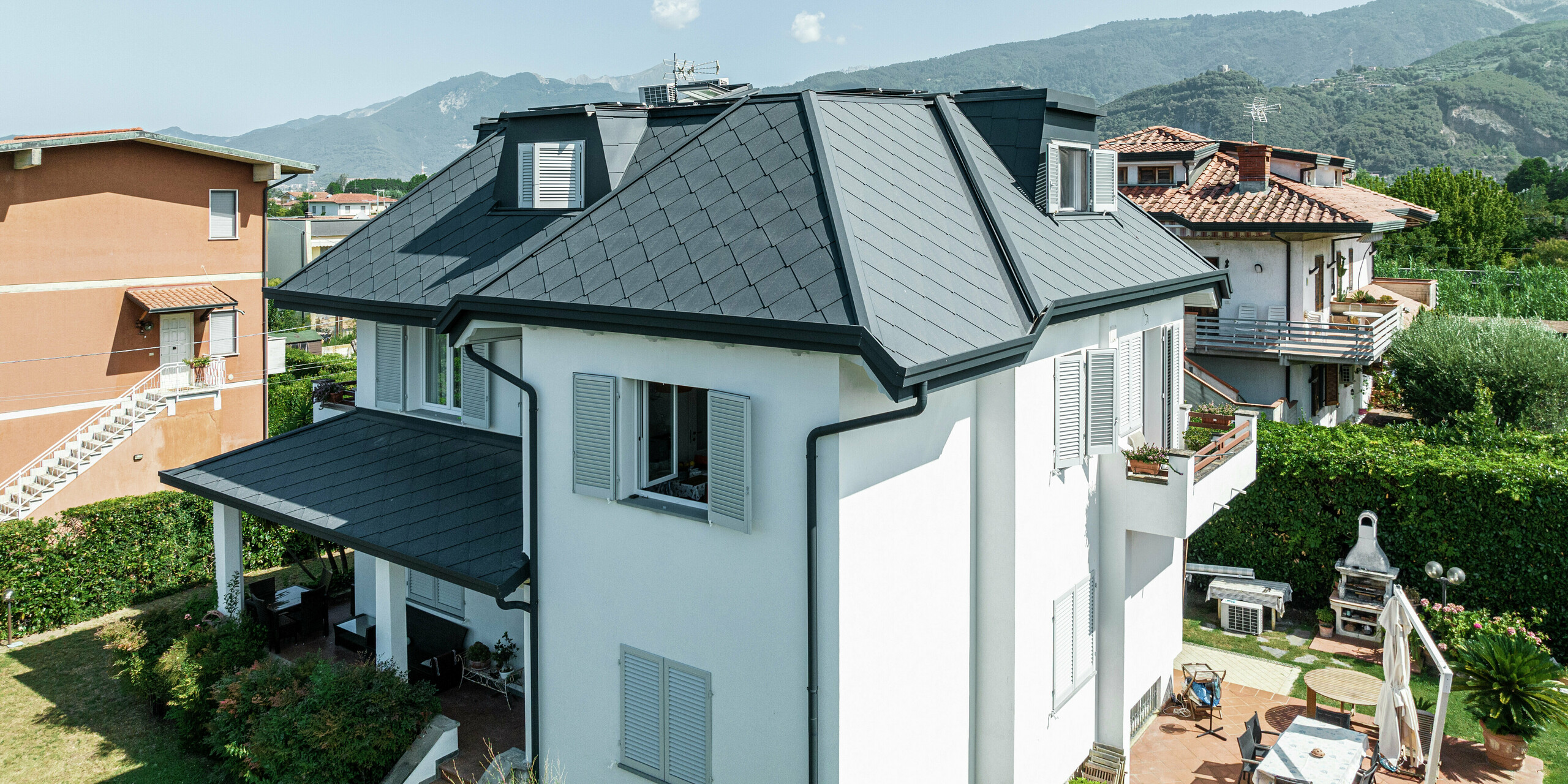 Nahaufnahme des eleganten Daches eines Einfamilienhauses in Italien, eingedeckt mit der PREFA Dachraute 44 × 44 in P.10 Steingrau. Die klare Linienführung und die hochwertige Aluminiumdeckung unterstreichen das moderne Design des Einfamilienhauses in Montignoso. Dank der robusten PREFA Materialien ist das Dach nicht nur optisch ansprechend, sondern auch extrem widerstandsfähig gegen Sturm, Korrosion und UV-Strahlung. Die dunkle Dacheindeckung bildet einen stilvollen Kontrast zur hellen Fassade und fügt sich harmonisch in die Umgebung ein.