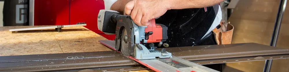 Das Bild zeigt einen Mann, der an einer Werkbank arbeitet. Er schneidet mit einer Sägemaschine das PREFA Siding-Fassadenelement zu. Er ist dabei sehr fokussiert, die Detailgenauigkeit seiner Arbeit kommt zum Vorschein.