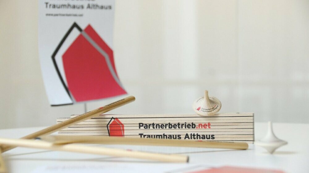 Zollstock mit Aufdruck „Partnerbetrieb.net Traumhaus Althaus“ auf einem Tisch, umgeben von Holzstäben und einem Kreisel, im Hintergrund ein Schild mit Haus-Logo.