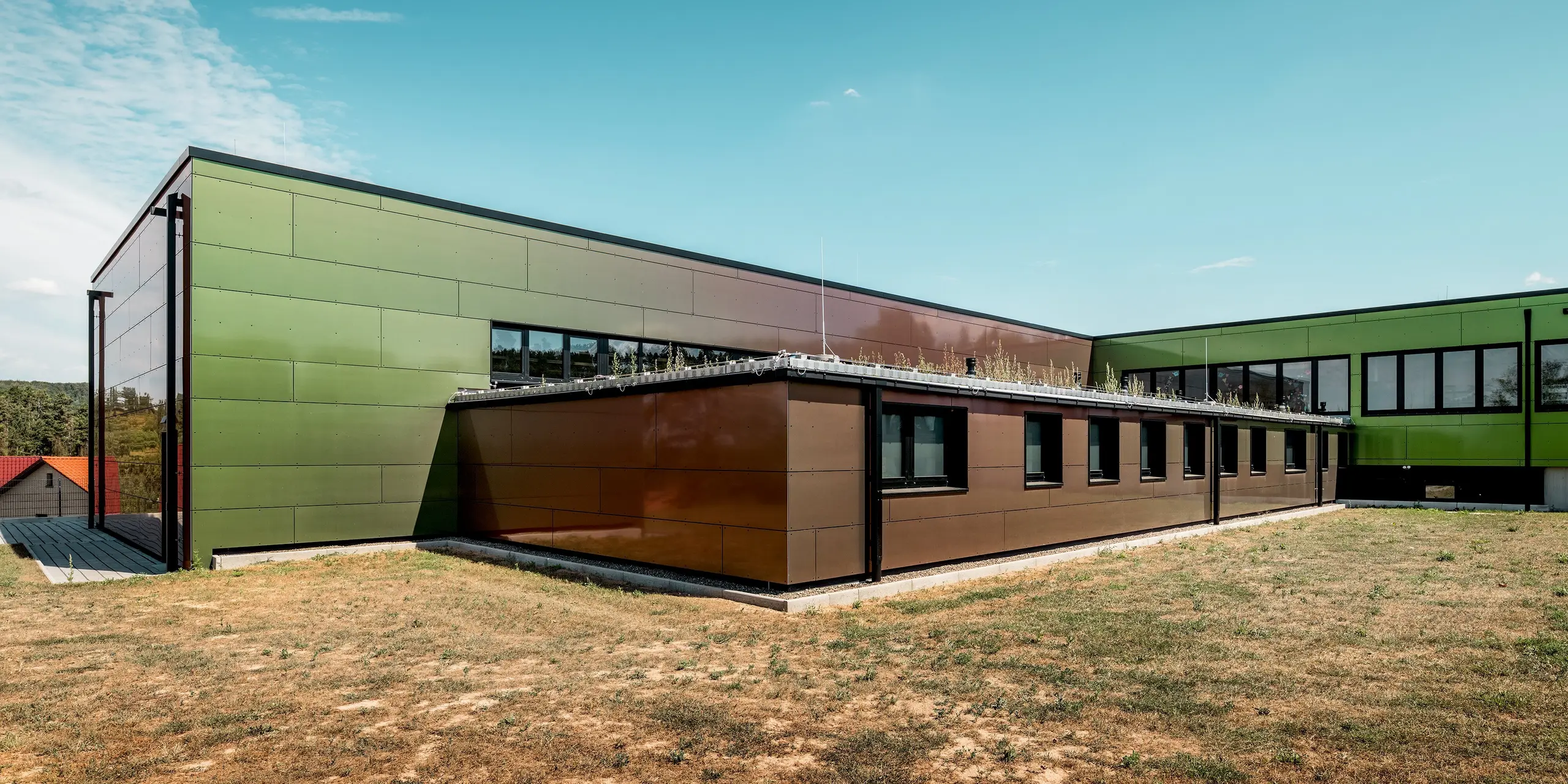 Die moderne Schularchitektur der Johannwolfschule in Angerstein zeichnet sich durch unterschiedlich hohe Gebäudetrakte mit Flachdächern und langen Fensterreihen aus. Die großformatigen PREFA Aluminium Verbundplatten in der Sonderfarbe „Spring Lake” schimmern je nach Lichteinfall in Grün- und Bronzetönen an der Fassade. Ein begrüntes Flachdach und eine dezente Dachentwässerung mit PREFA Quadratrohren ergänzen die geradlinige Architektur.
