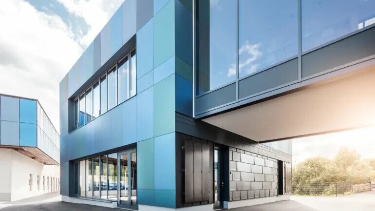 Moderner Gebäudekomplex mit blauer Aluminiumfassade, großen Glasflächen und klaren Linien, aufgenommen aus der Perspektive einer Gebäudeecke mit Durchfahrt.