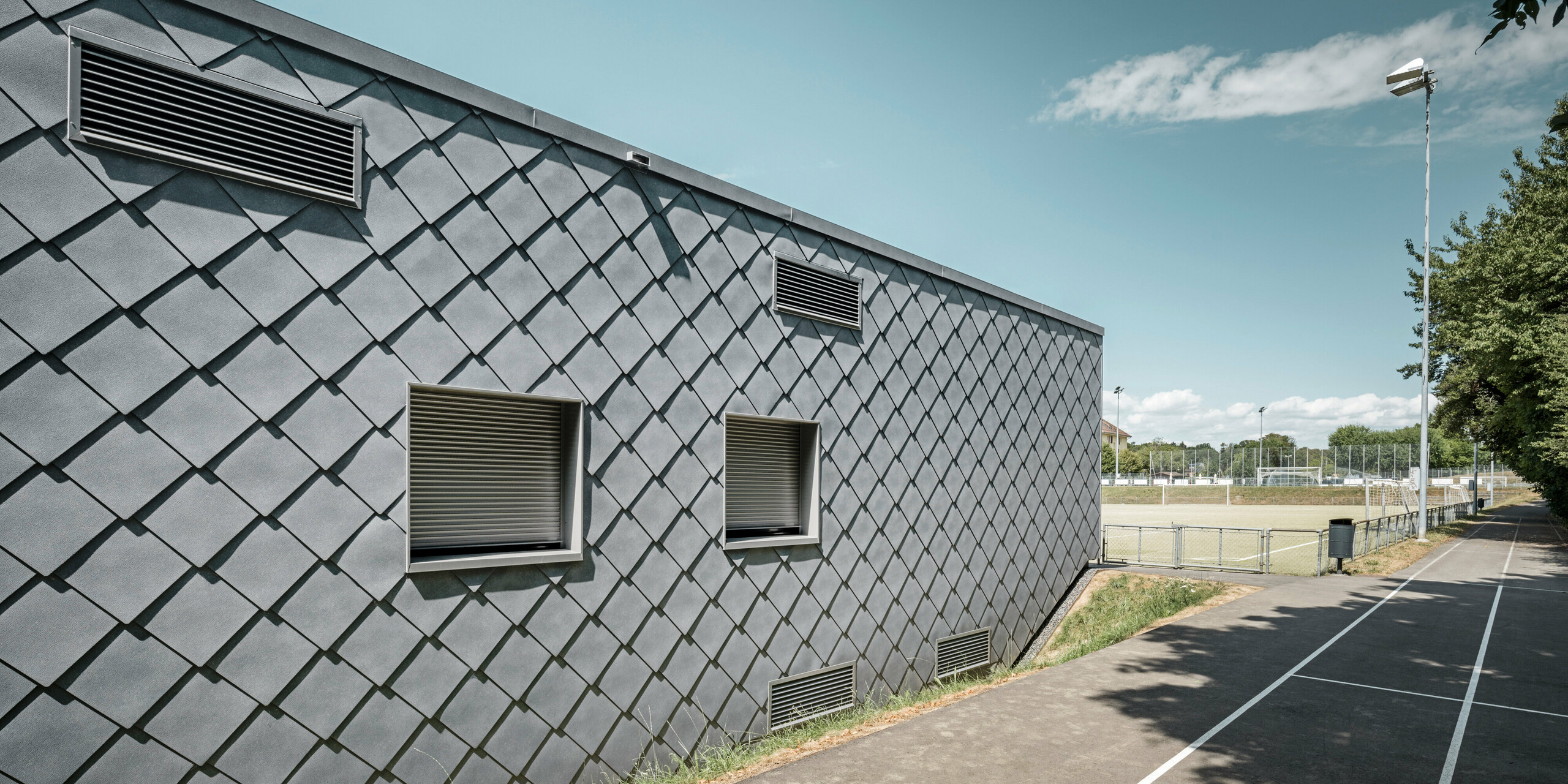 Rückseite einer Sportanlage in Prilly, Schweiz, mit einer robusten Aluminiumhülle aus PREFA Dach- und Wandrauten 44x44 in P.10 Steingrau. Die Detailaufnahme zeigt die präzise Verarbeitung und hochwertige Qualität der kleinformatigen Aluminiumprodukte, die Langlebigkeit und Witterungsbeständigkeit gewährleisten. Am Gebäude sind mehrere Lüftungsgitter in unterschiedlichen Formen und Größen installiert worden. Die Wandrauten an der Fassade sorgen für eine ästhetische Erscheinung des Gebäudes und fügen sich nahtlos in die sportliche Umgebung ein, neben einem Fußballfeld und einer Laufbahn. Im Hintergrund sind mehrere Flutlichtmasten zu sehen, die das Fußballfeld bei Nacht beleuchten.