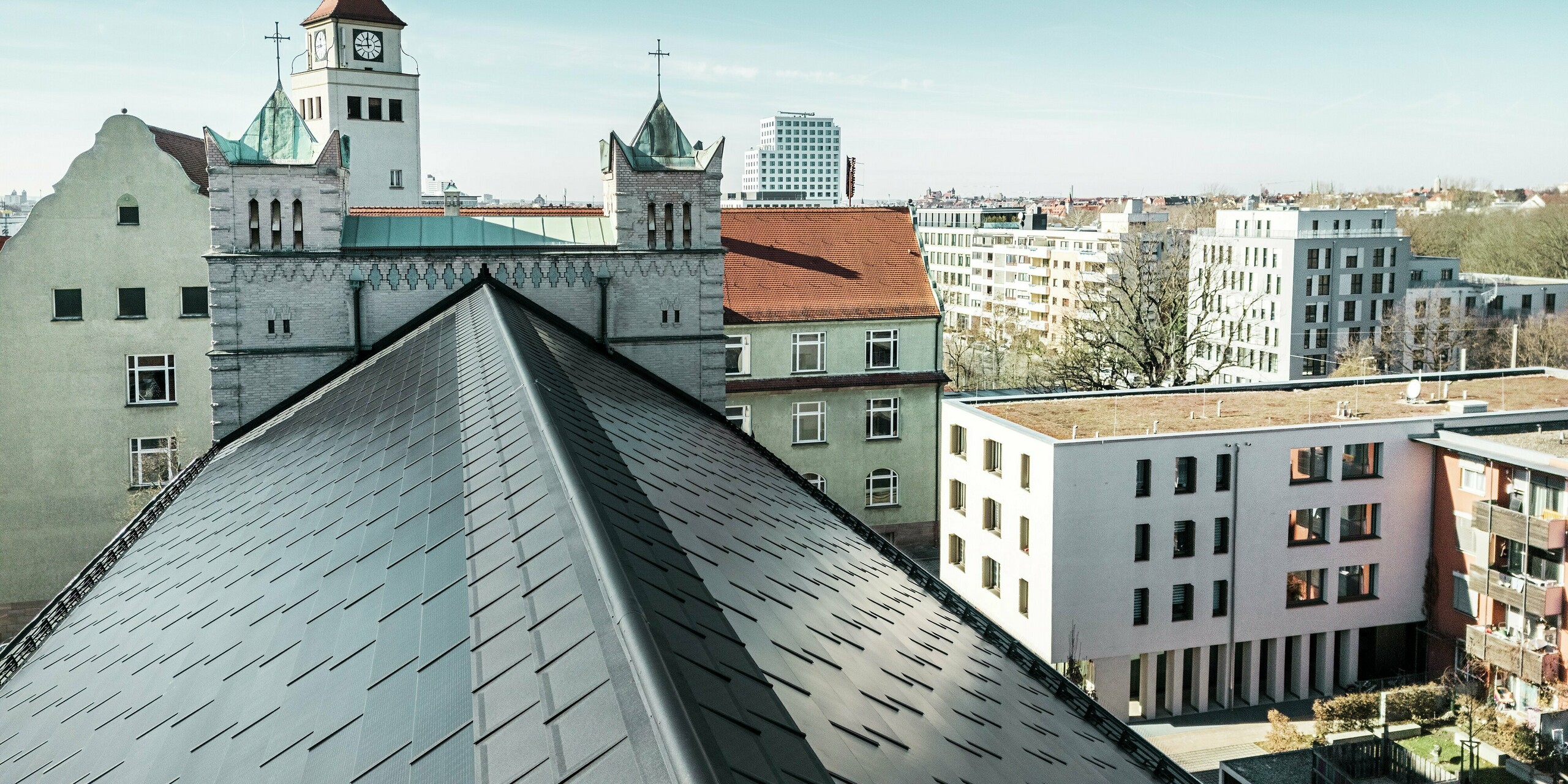 Das Bild zeigt die präzise Deckung des Aluminiumdachs der St. Karl Borromäus Kirche in Nürnberg. Das Kirchendach wurde mit PREFA Solardachplatten in elegantem Schwarz ausgestattet. Der Blick auf den markanten First des Satteldachs unterstreicht die gelungene Integration der modernen Photovoltaik-Indach-Lösung in die historische Architektur. Die Solardachplatten fügen sich harmonisch auf dem denkmalgeschützten Gebäude ein und versorgen die Kirche und etliche benachbarte Gebäude mit effizienter und nachhaltiger Energie.
