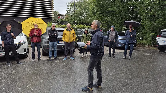 Eine Gruppe von Architekturschaffenden und Fachleuten verfolgt trotz Regen aufmerksam eine Präsentation vor einem markanten Firmengebäude. Eyecatcher ist die moderne Fassadengestaltung mit horizontalen Lamellen. Regenschirme setzen lebendige Akzente im Bild.