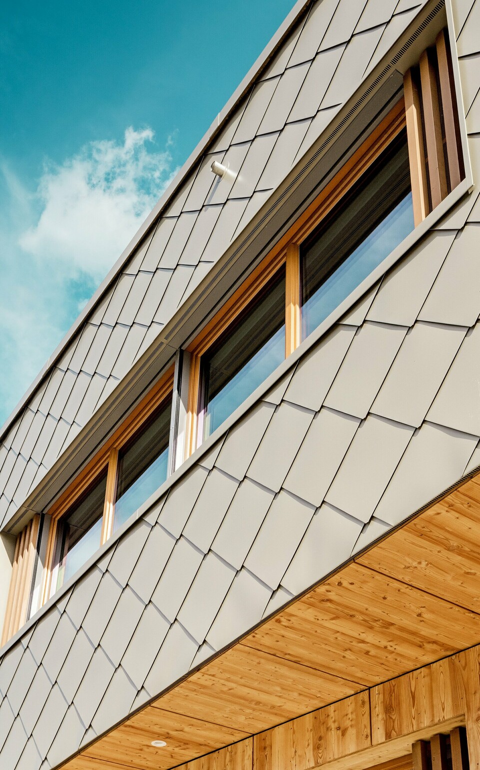 Die Nahaufnahme der Kindertagesstätte in Alzenau zeigt die präzise Ausführung und hochwertige Materialkombination aus bronzefarbenen PREFA Wandrauten 44 × 44 und naturbelassenem Holz. Die diagonale Struktur der Aluminiumfassade bildet ein markantes Schuppenmuster, das sich harmonisch mit den vertikalen Holzlamellen an den Fenstern und der fein ausgearbeiteten Holzuntersicht verbindet. Die Aufnahme aus der Froschperspektive betont die exakte Linienführung und handwerkliche Qualität der vorgehängten hinterlüfteten Fassade. Diese Detailansicht unterstreicht die gestalterische Stärke und Langlebigkeit von PREFA Produkten – ideal für nachhaltige Architekturprojekte, die Ästhetik, Funktionalität und Ökologie auf höchstem Niveau vereinen.