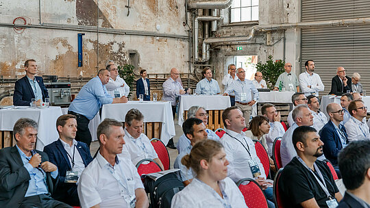 Aufmerksames Fachpublikum beim Forum Aluminium in einer industriellen Eventlocation: Zahlreiche Teilnehmende sitzen auf roten Stühlen oder stehen an Stehtischen und verfolgen konzentriert die Präsentation. Die raue, historische Architektur mit freiliegenden Rohren, Betonwänden und großen Fenstern bildet einen markanten Kontrast zur modernen Business-Atmosphäre. Namensbadges, Getränke und Informationsmaterialien unterstreichen den professionellen Networking-Charakter und zeigen den intensiven Austausch zu Innovationen, Recycling und Zukunftsthemen der Aluminiumindustrie.
