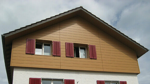 Giebelverkleidung eines klassischen Hauses mit Satteldach. Der Giebel ist mit horizontal verlegten PREFA Sidings in Eiche natur verkleidet. Die Fenster haben rote Fensterläden.
