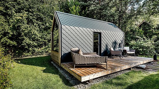 Ein minimalistisches Tiny House im Secret Koi Garden am Natterer See steht auf einer großzügigen Holzterrasse, umgeben von dichter Waldvegetation. Dach und Fassade sind mit kleinteiligen PREFA Dach‑ und Wandrauten aus Aluminium in P.10 Anthrazit ausgeführt und prägen mit ihrer gleichmäßigen Struktur das moderne Erscheinungsbild. Die langlebige, witterungsbeständige Aluminiumhülle schützt zuverlässig und bildet einen starken Kontrast zu Holz, Grünfläche und dem offenen Outdoor‑Wohnbereich des Glamping‑Resorts.