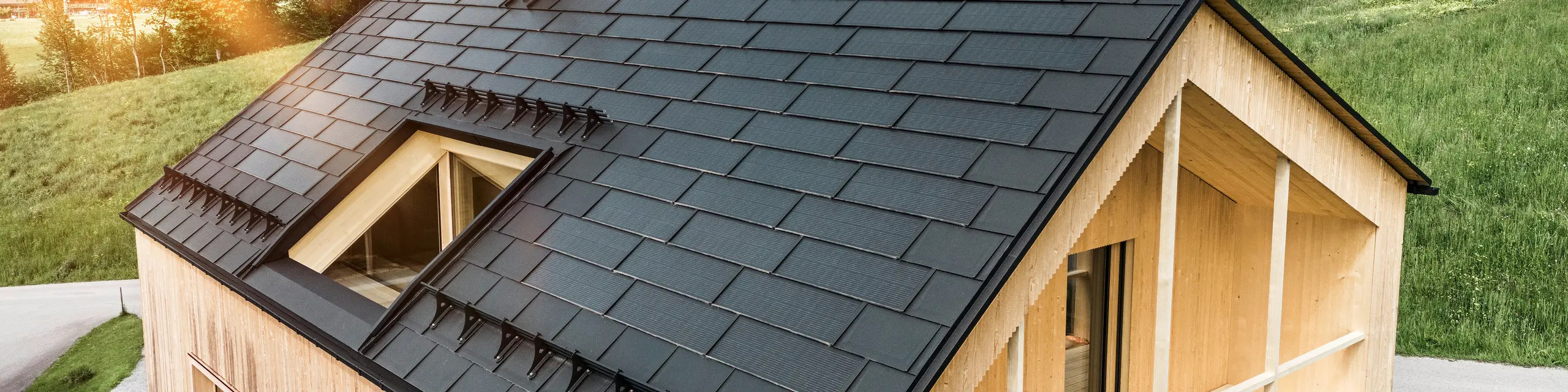 Blick auf ein modernes, nachhaltiges Holzhaus in Egg mit einem PREFA Solardach, das durch seine dunkle, strukturierte sowie elegante Erscheinung auffällt. Die Solarmodule sind strategisch zwischen den klassischen Dachplatten R.16 platziert, um eine optimale Sonneneinstrahlung zu nutzen und sich dabei ästhetisch in das Gesamtdesign einzufügen. Das Haus steht im Einklang mit seiner grünen Umgebung, während die warme Abendsonne die natürliche Holztextur der Fassade unterstreicht und die solide Bauweise betont. Ein harmonisches Beispiel für umweltbewusstes Bauen und Energieeffizienz.