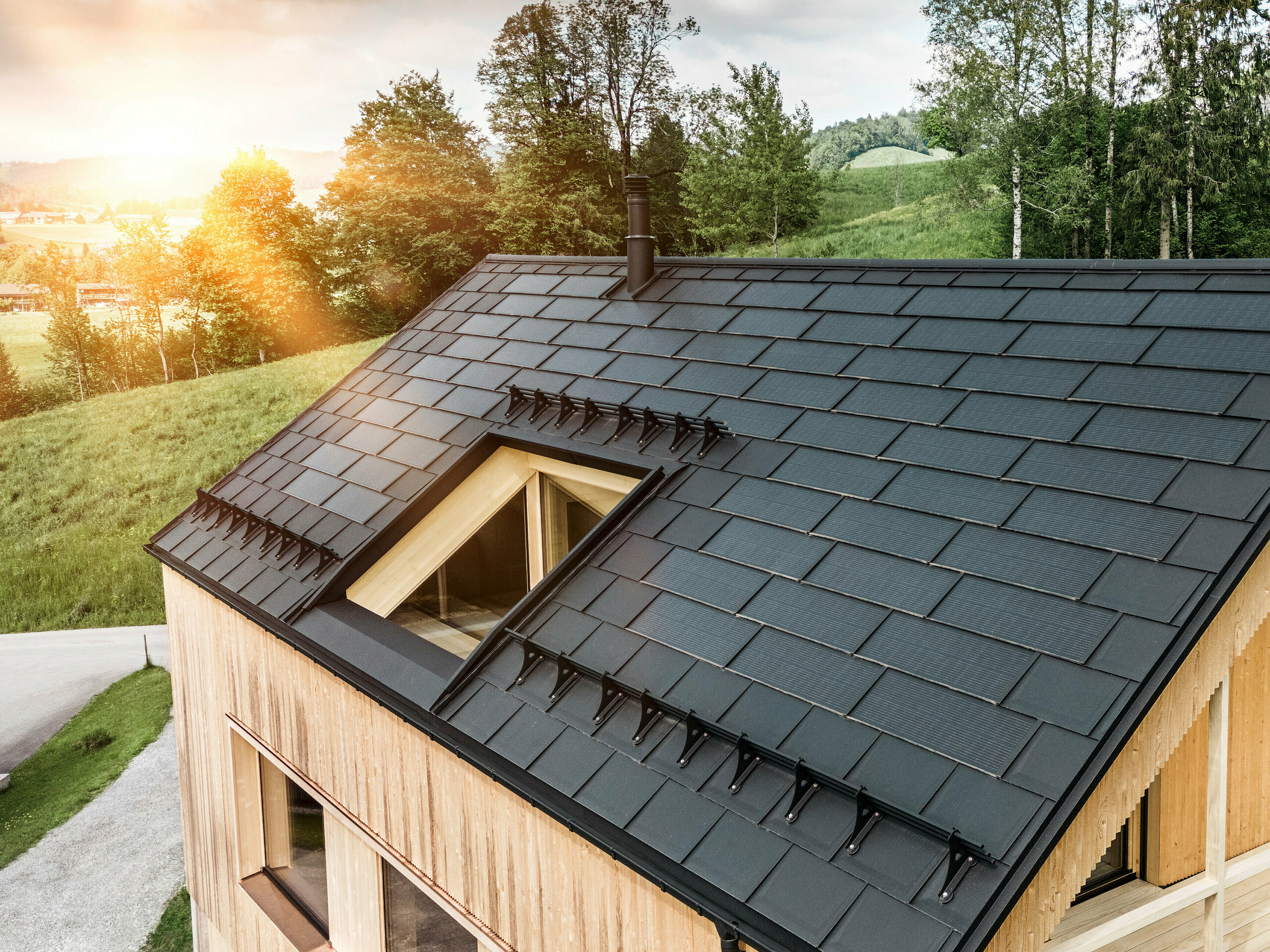 Blick auf ein modernes, nachhaltiges Holzhaus in Egg mit einem PREFA Solardach, das durch seine dunkle, strukturierte sowie elegante Erscheinung auffällt. Die Solarmodule sind strategisch zwischen den klassischen Dachplatten R.16 platziert, um eine optimale Sonneneinstrahlung zu nutzen und sich dabei ästhetisch in das Gesamtdesign einzufügen. Das Haus steht im Einklang mit seiner grünen Umgebung, während die warme Abendsonne die natürliche Holztextur der Fassade unterstreicht und die solide Bauweise betont. Ein harmonisches Beispiel für umweltbewusstes Bauen und Energieeffizienz.