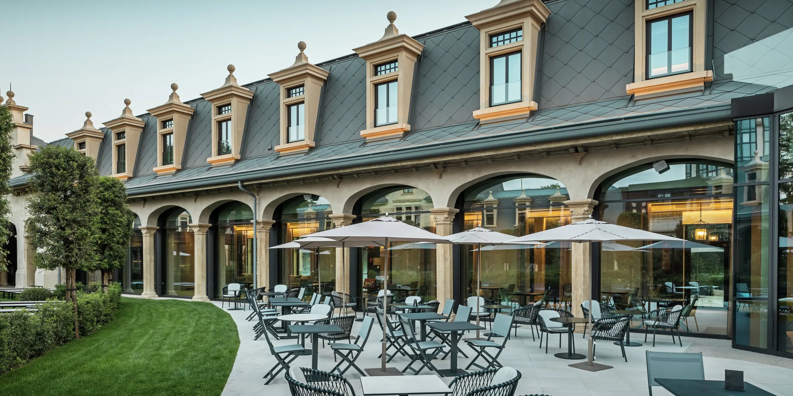 Die stilvoll gestaltete Terrasse des Thermalparks „De Montel – Terme Milano“ lädt mit modernen Sitzgruppen und Sonnenschirmen zum Verweilen ein. Im Hintergrund trifft denkmalgeschützte Architektur auf moderne Funktionalität: Das Aluminiumdach mit PREFA Dachrauten 44 × 44 in P.10 Hellgrau fügt sich harmonisch in die fein restaurierte Jugendstilfassade ein. Großflächige Glasfronten schaffen Transparenz und verbinden Innen- und Außenraum auf elegante Weise.