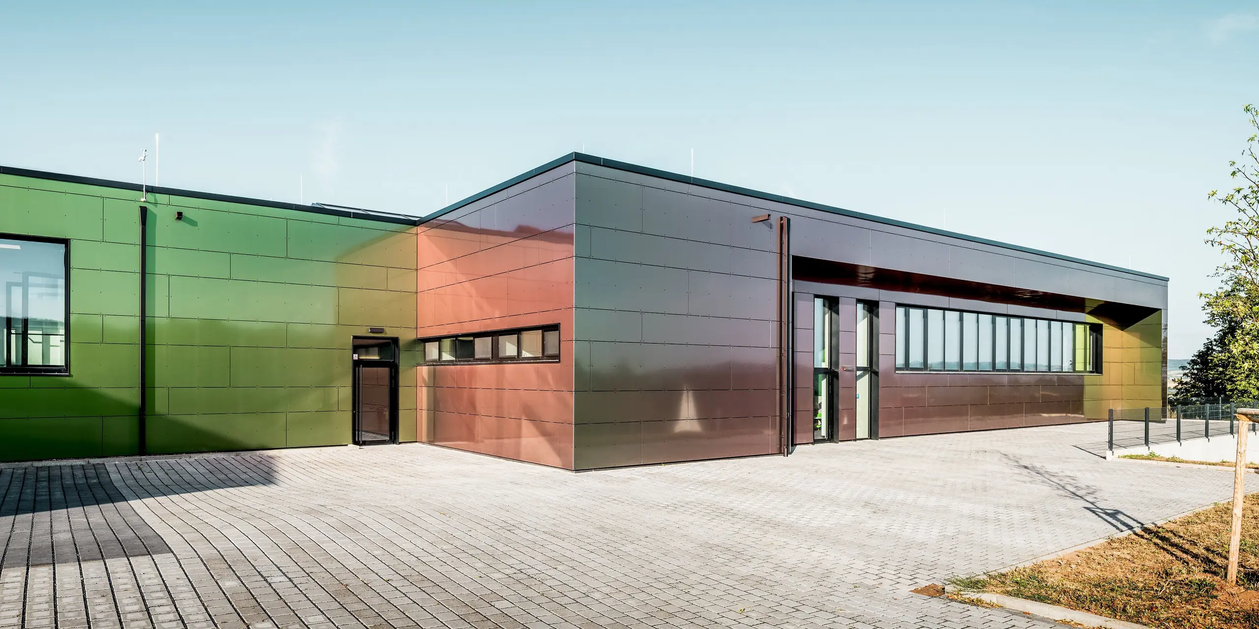 Moderne Architektur der Johannwolfschule Angerstein: Zwei geradlinige Gebäudetrakte mit großzügigen Fensterfronten und gepflastertem Vorplatz beeindrucken durch ihre außergewöhnliche PREFA Fassade. Die großformatigen Aluminium Verbundplatten in der Sonderfarbe Spring Lake schimmern im Sonnenlicht in lebendigen Grün-, Bronze- und Rottönen. Dunkle PREFA Quadratrohre ergänzen die langlebige Fassadengestaltung harmonisch.