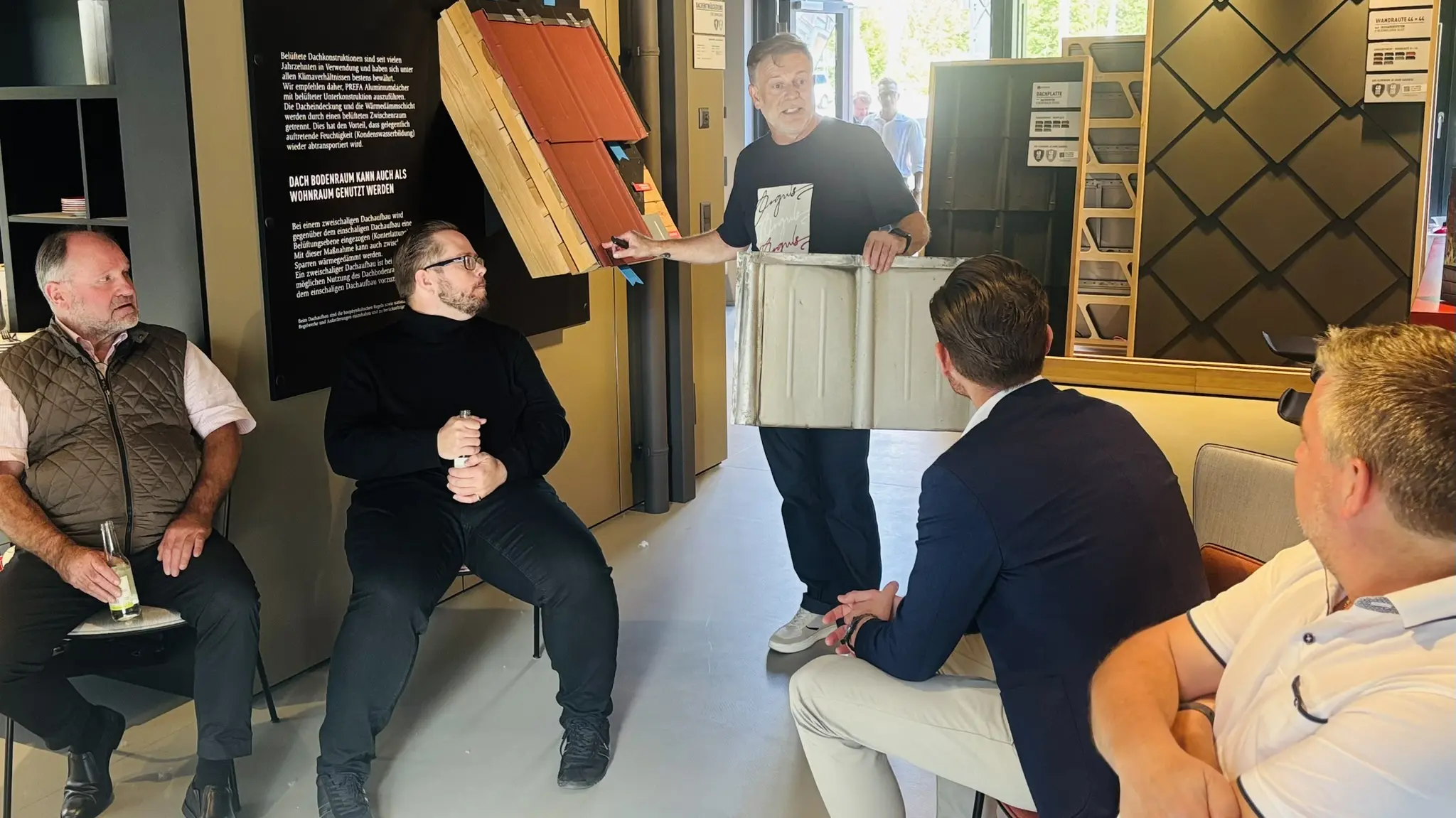 Teilnehmende des PREFA Barcamp-Abends im Showroom St. Veit an der Glan verfolgen eine praxisnahe Präsentation zum Dachaufbau mit PREFA Aluminiumlösungen. Ralf Stadlober, PREFA Objektberater, erläutert anhand eines Musterquerschnitts und einer ausgestellten PREFA Dachplatte den mehrschichtigen Aufbau von Dachsystemen. Im Hintergrund sind Infotafeln zum zweischaligen Dachaufbau sowie Fassadenmuster sichtbar. Die Szene zeigt intensiven fachlichen Austausch, anschauliche Produktdemonstration und vermittelt praxisnahes Know-how für die Planung moderner PREFA Dach- und Fassadenprojekte.