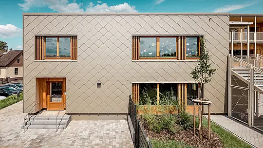 Die Rückseite der modernen Kindertagesstätte in Alzenau wird aus einer frontalen Perspektive gezeigt und beeindruckt mit klaren Linien und einer markanten Fassade aus bronzefarbenen PREFA Wandrauten 44 × 44. Die geometrische Fassadenstruktur erzeugt ein harmonisches Schuppenmuster, das durch die ruhige, horizontale Ausrichtung der Fenster akzentuiert wird. Großzügige Holzrahmen und vertikale Lamellen setzen natürliche Akzente und sorgen für eine warme, einladende Atmosphäre. Die durchdachte Kombination aus langlebigem Aluminium und regionalem Holz steht für nachhaltige Architektur mit hoher gestalterischer Qualität. Das Bild betont die präzise Ausführung, die hochwertige Materialwahl und die gelungene Integration des Gebäudes in das städtische Umfeld.