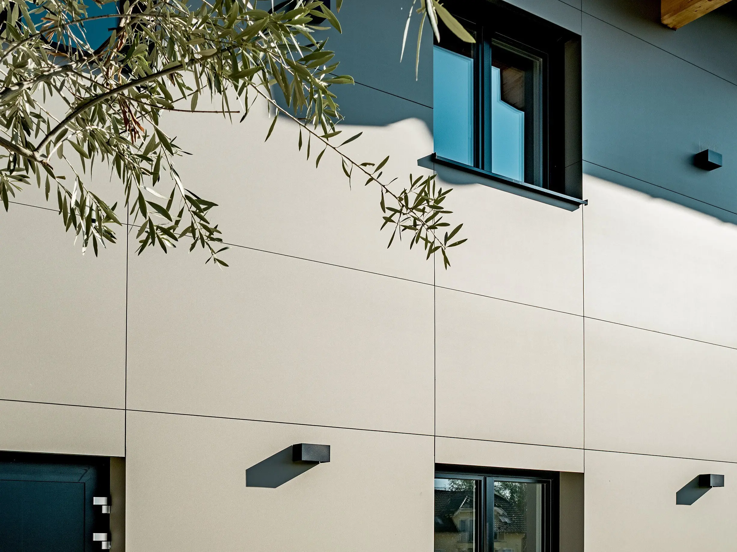 Nahaufnahme der Fassade des Weinguts Nigl in Perchtoldsdorf mit PREFA Aluminium Verbundplatten in P.10 Prefabronze. Die glatte, matte Oberfläche betont die hochwertige Verarbeitung und verleiht dem Gebäude eine moderne, zurückhaltende Eleganz. Klar definierte Fugenlinien sorgen für eine strukturierte Optik. Im Vordergrund steht ein alter Olivenbaum, dessen natürliche Maserung einen reizvollen Kontrast zur präzisen Linienführung der Fassade bildet. Schwarze Fensterrahmen und reduzierte Außenleuchten runden das minimalistische Design ab. Die PREFA Fassadenplatten sind witterungsbeständig, wartungsarm und besonders langlebig – ideal für zeitlose Architektur mit Stil.