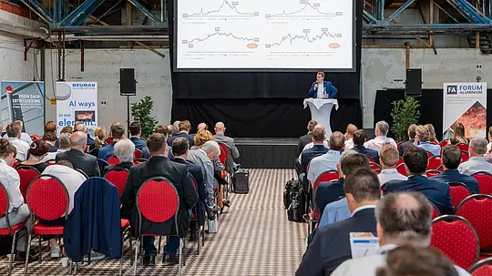 Großer Konferenzsaal im industriellen Ambiente des Glanzstoff-Geländes mit dicht besetzten Reihen und roter Bestuhlung als visuellem Kontrast. Im Fokus steht eine beleuchtete Bühne mit Rednerpult und Präsentationsfläche, auf der detaillierte Marktanalysen zu Rohstoff- und Energiepreisen gezeigt werden. Flankiert von Ausstellerbannern wie PREFA Dachentwässerung und Neuman Aluminium entsteht eine dynamische Atmosphäre. Das Bild vermittelt den intensiven Austausch führender Experten beim Forum Aluminium zu Innovationen, Markttrends und Zukunft der Branche.