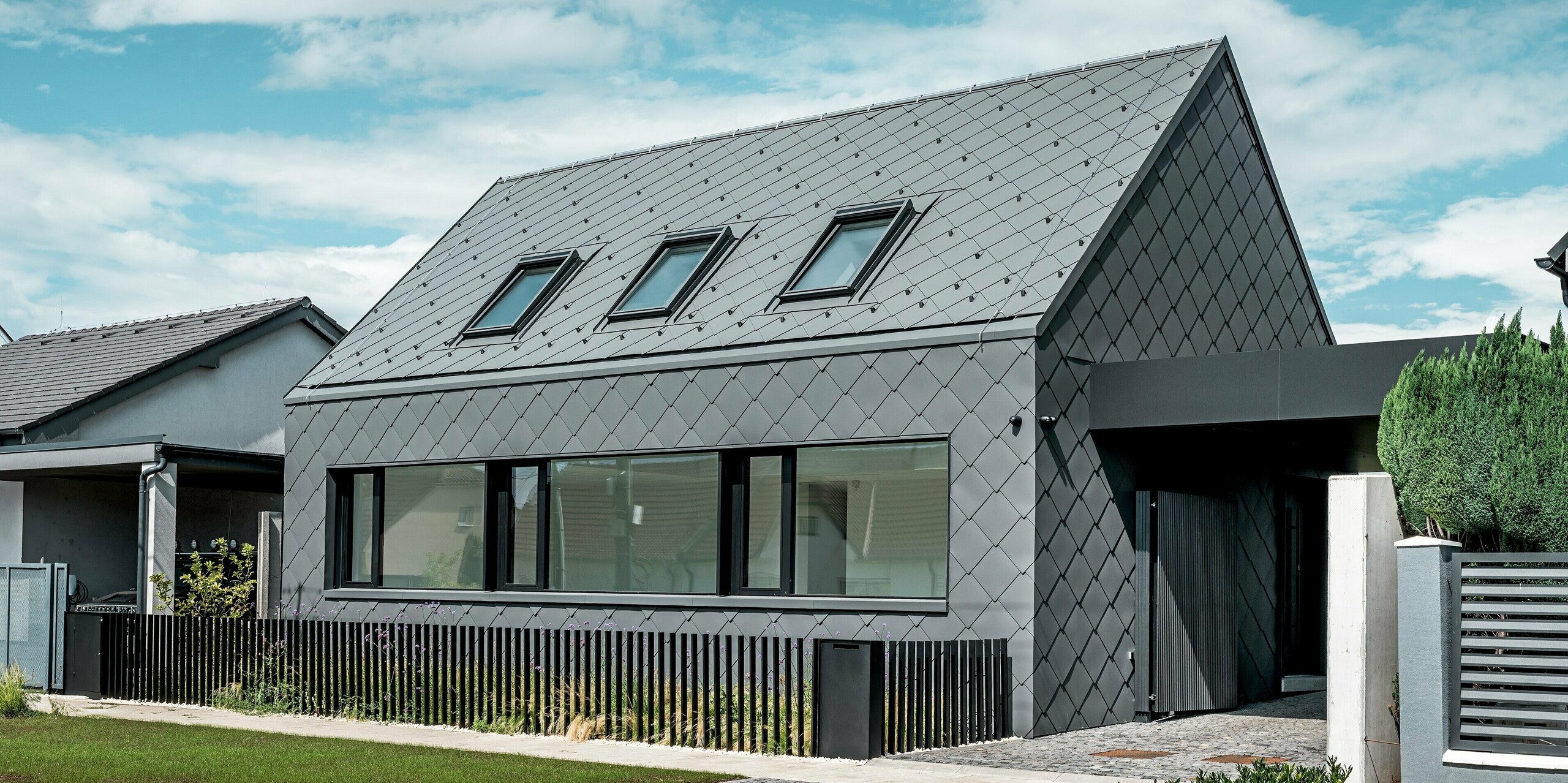 Modernes Einfamilienhaus in Trnava‑Modranka mit Aluminiumhülle: Die gesamte Gebäudehülle dieses schlichten Gebäudes wurde mit PREFA Dach- und Wandrauten 44 × 44 in P.10 Dunkelgrau gestaltet. Die markante Rautenstruktur zieht sich nahtlos über Dach und Fassade und verleiht dem Haus eine starke, monolithische Wirkung. Große Fensteröffnungen setzen klare architektonische Akzente, während das witterungsbeständige PREFA Aluminium langlebigen Schutz und eine edle, matte Optik bietet. Der helle Himmel verstärkt den Kontrast zur dunklen Gebäudehülle und betont das moderne Design.