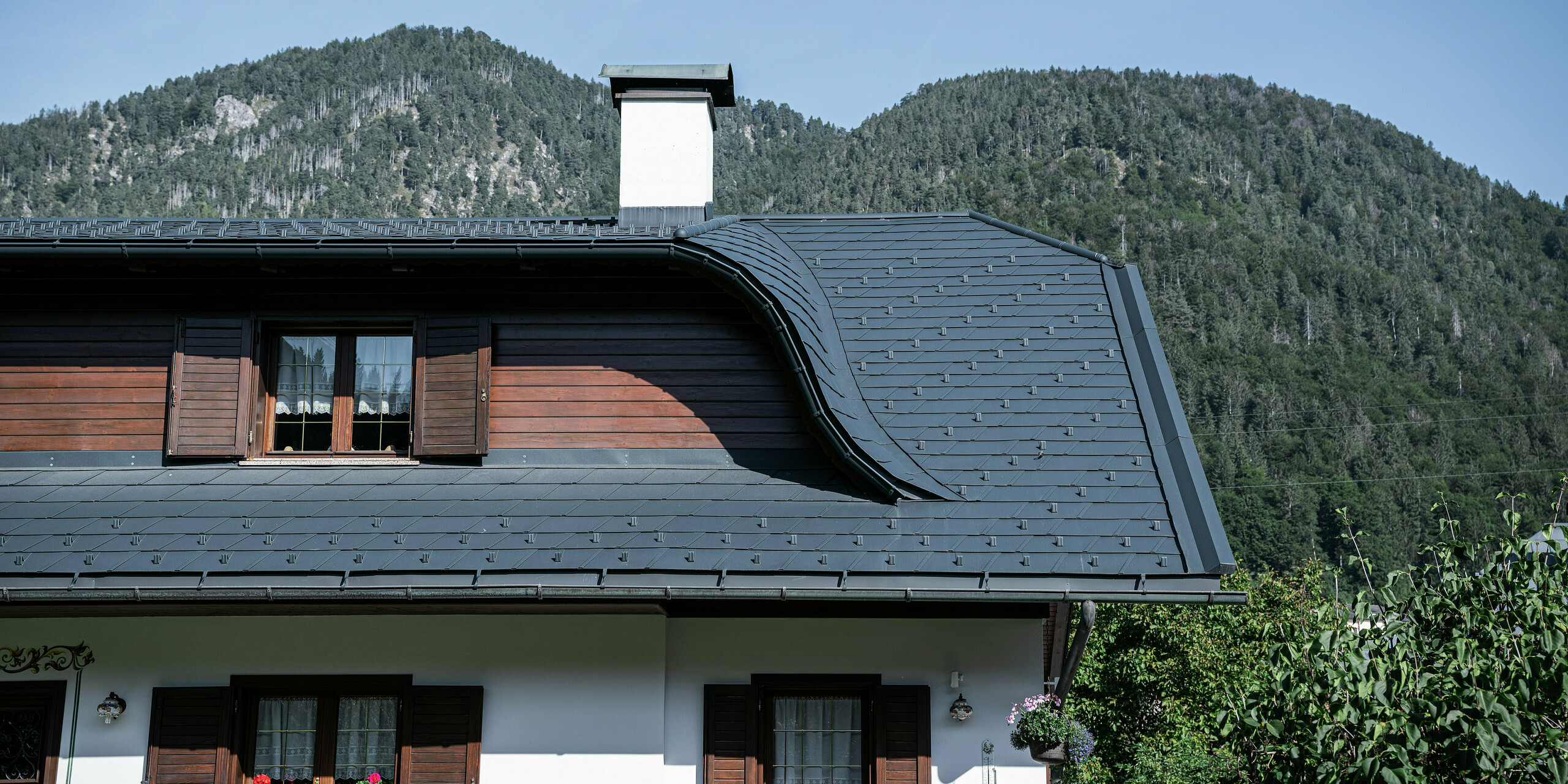 Nahaufnahme des Daches eines Einfamilienhauses in Tarvis, Italien, nach der Sanierung mit PREFA Dachschindeln P.10 anthrazit. Die elegante Hechtgaube verleiht dem Dach seine geschwungene Form und unterstreicht den traditionellen Stil des Hauses. Die langlebige Aluminium-Dacheindeckung bietet optimalen Schutz vor extremen Witterungseinflüssen. In Kombination mit der weißen Fassade und den Holzelementen entsteht ein harmonisches, alpines Gesamtbild, das sich perfekt in die Berglandschaft einfügt.